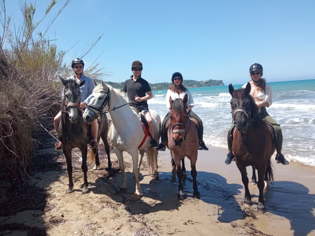 Four people riding horses on the beach near the shoreline with ocean waves and a small island or land formation in the background, wearing helmets.