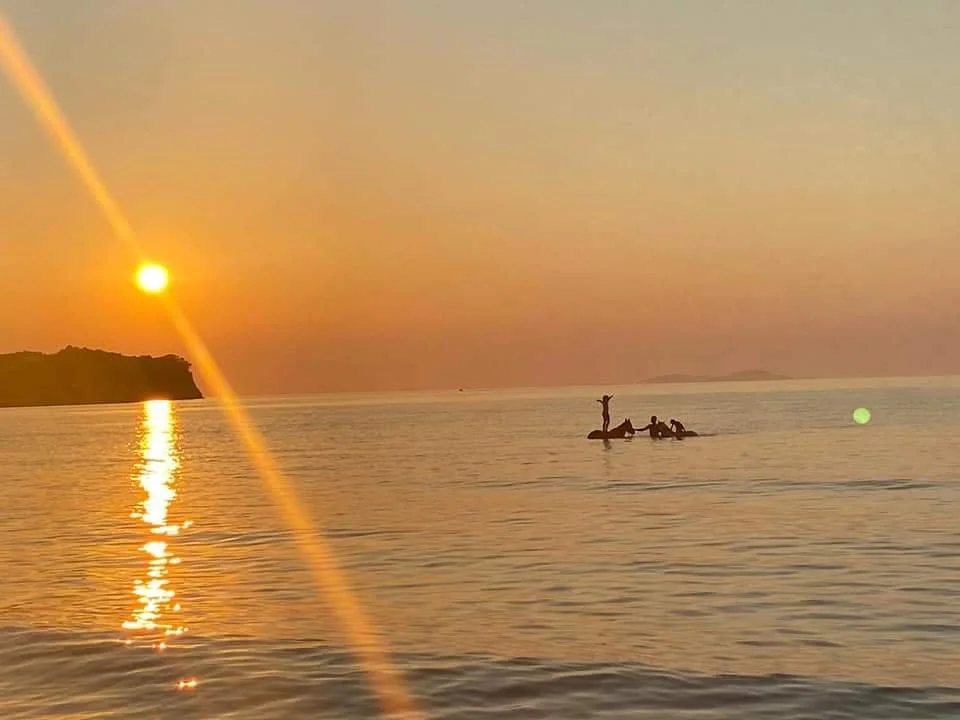 Sunset over calm water with sailboat silhouette and a small boat with two people on it.