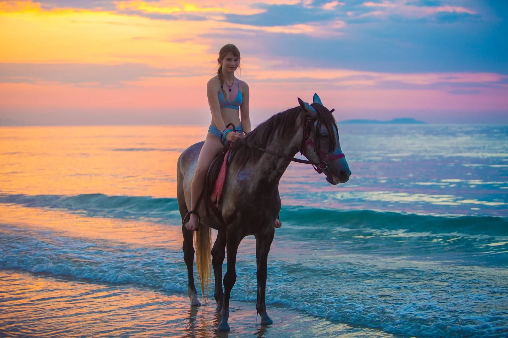 A girl riding a horse in the ocean during a colorful sunset.
