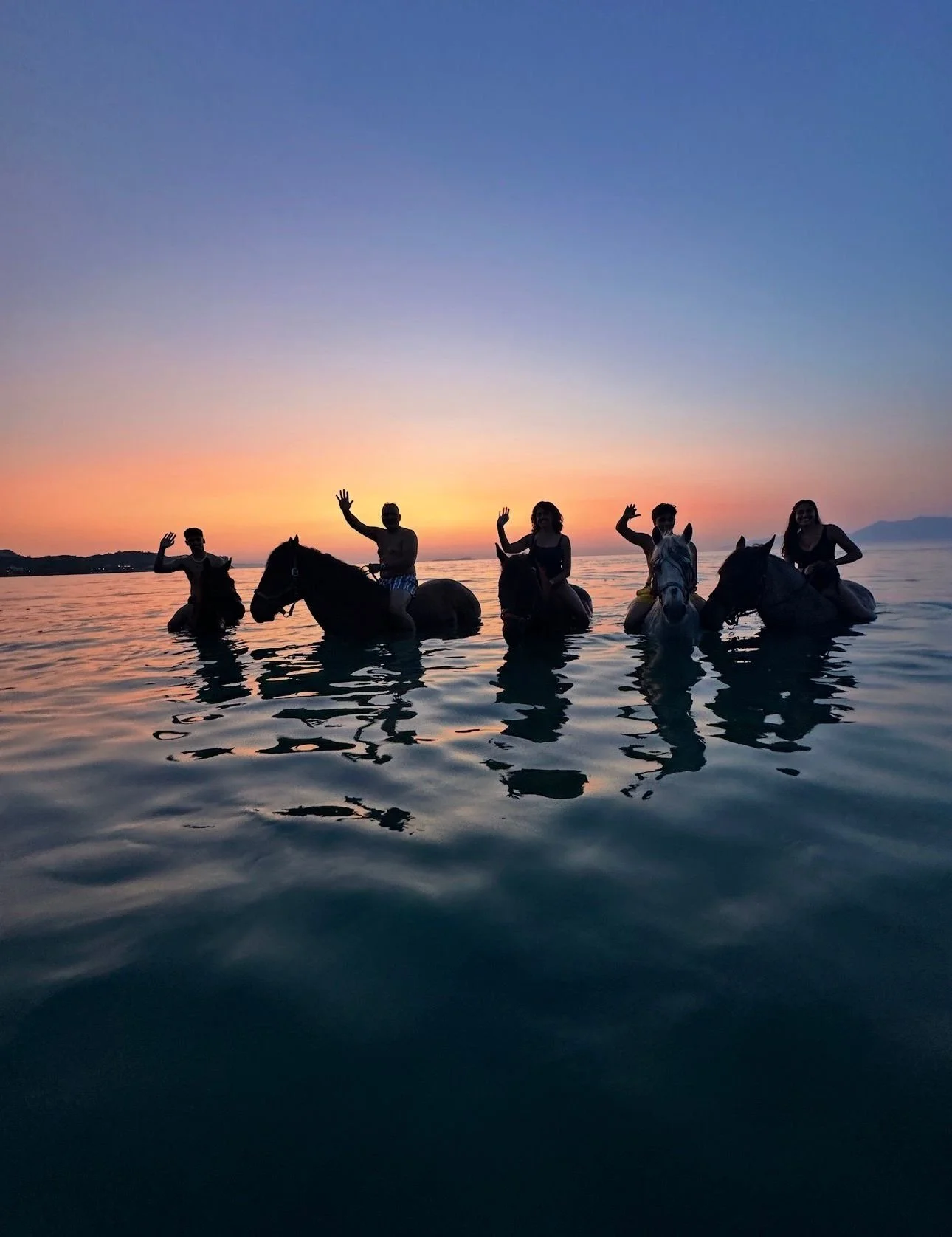 Group of five people riding horses in the water at sunset, waving, with a colorful sky in the background.