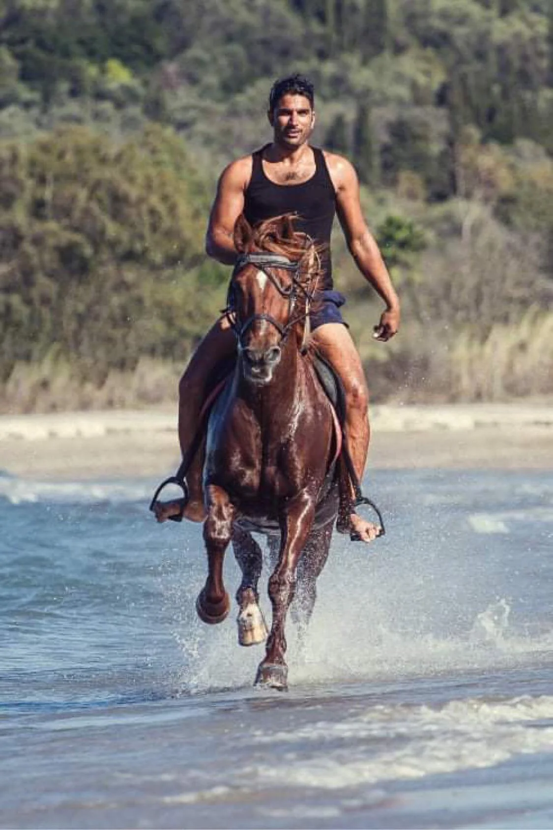 A man riding a horse through shallow water on a beach, with dense trees in the background.