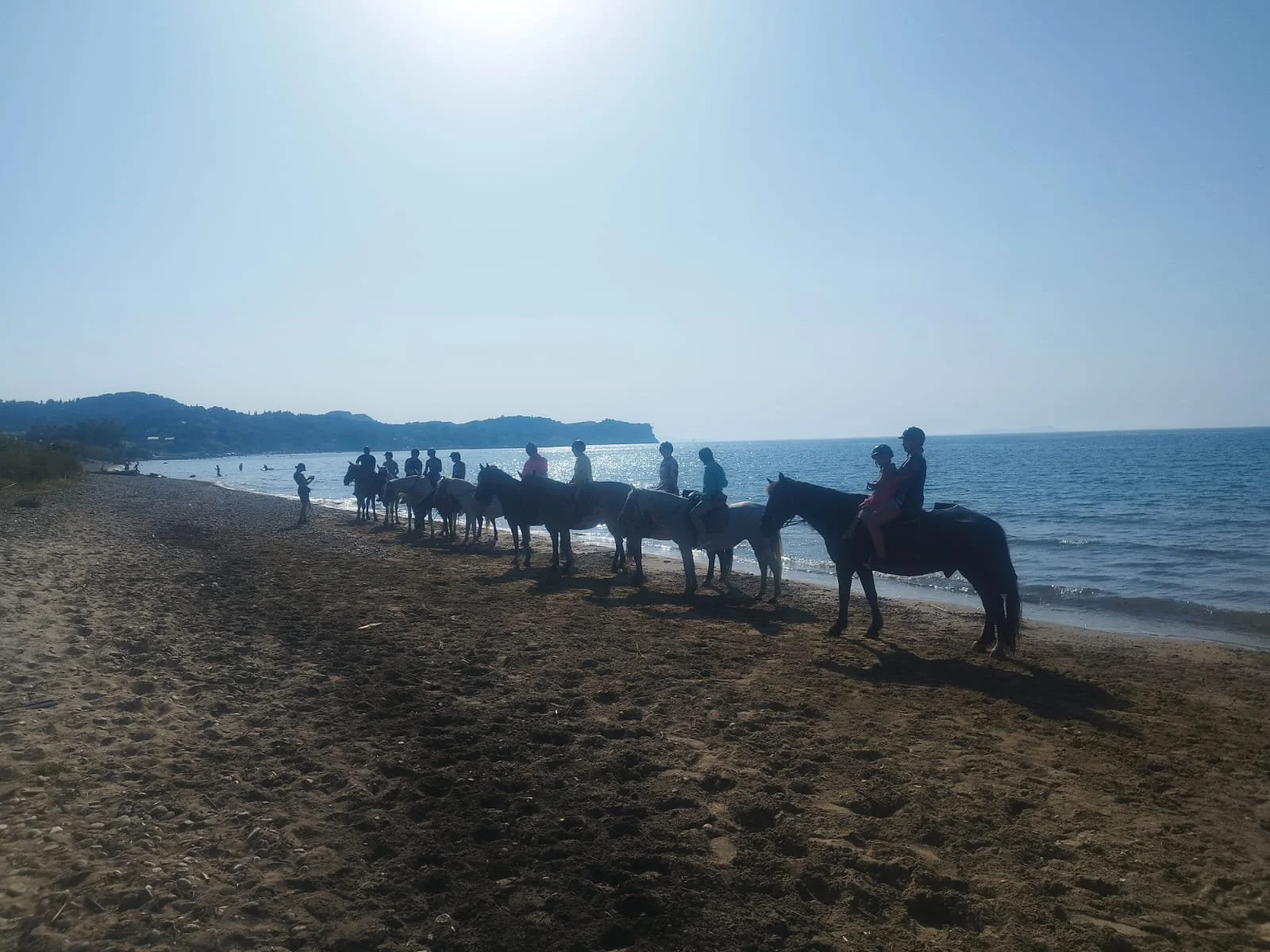 Group of people riding horses along the beach at sunset, with water and hills in the background.