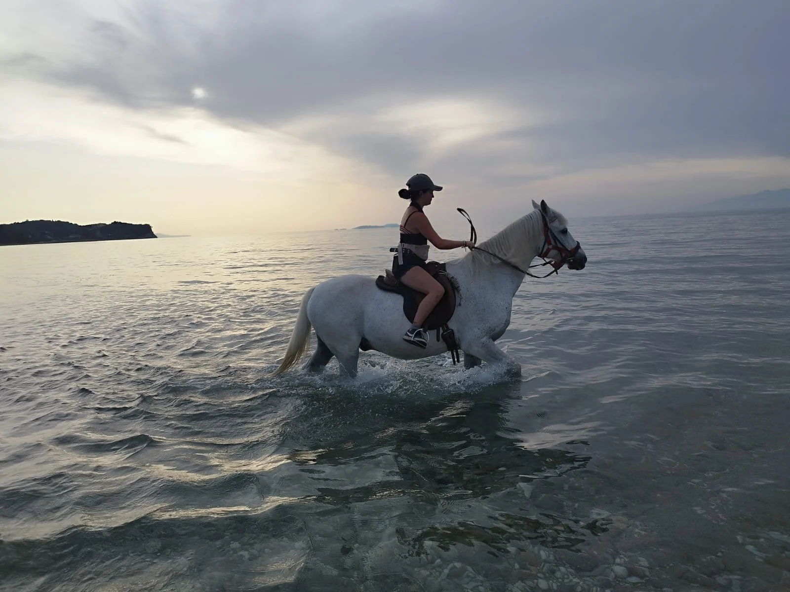 A woman riding a white horse through the shallow waters of a beach at sunset, with a cloudy sky and distant land in the background.