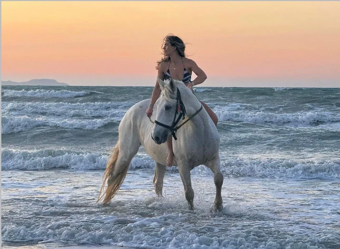 A woman riding a white horse in the ocean during sunset, with waves and a distant shoreline in the background.