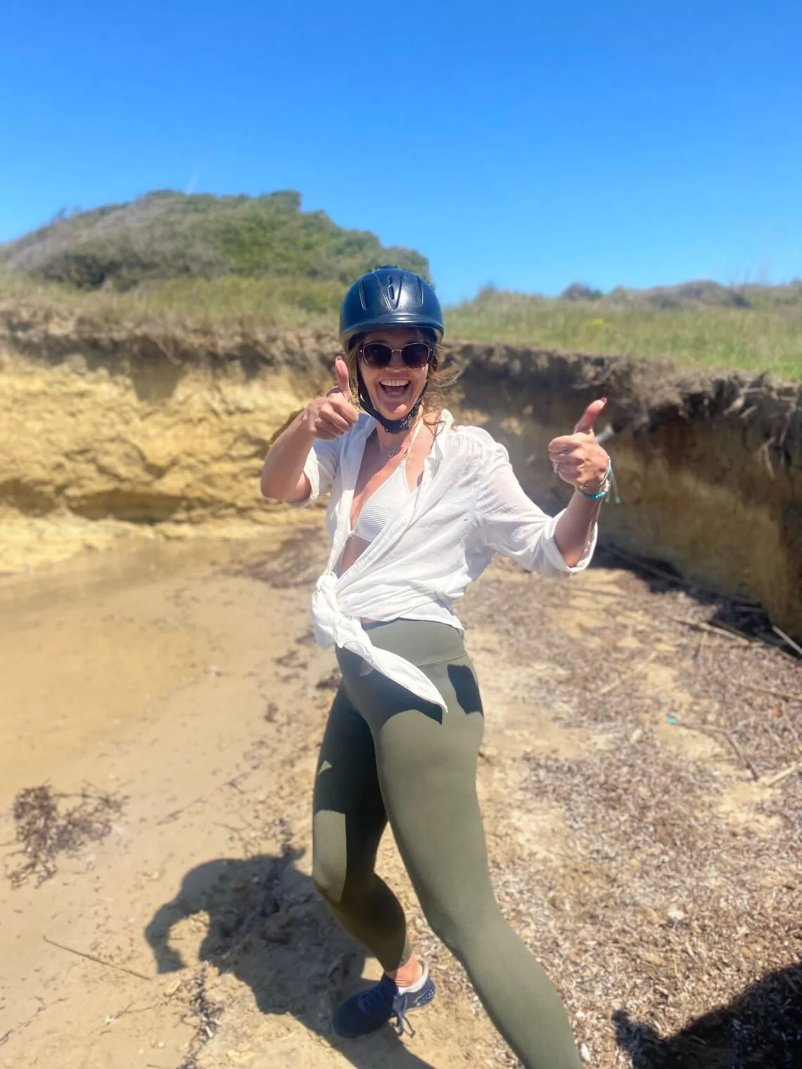 Woman wearing a black helmet and sunglasses, smiling and giving two thumbs up, standing outdoors on a sunny day with a sandy, rocky landscape and green hills in the background.