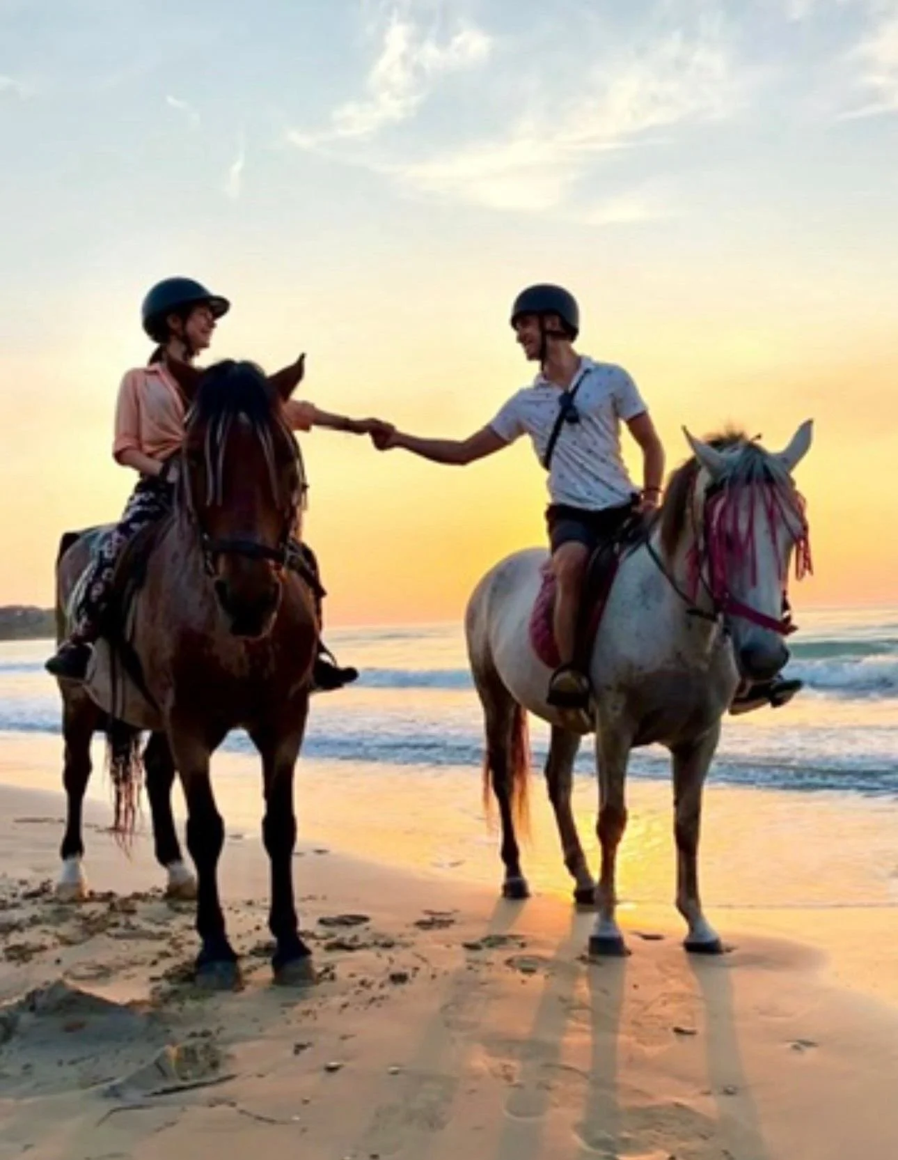 Two people riding horses on the beach at sunset, smiling and holding hands. Both are wearing helmets, and the sky is partly cloudy with a calm ocean in the background.