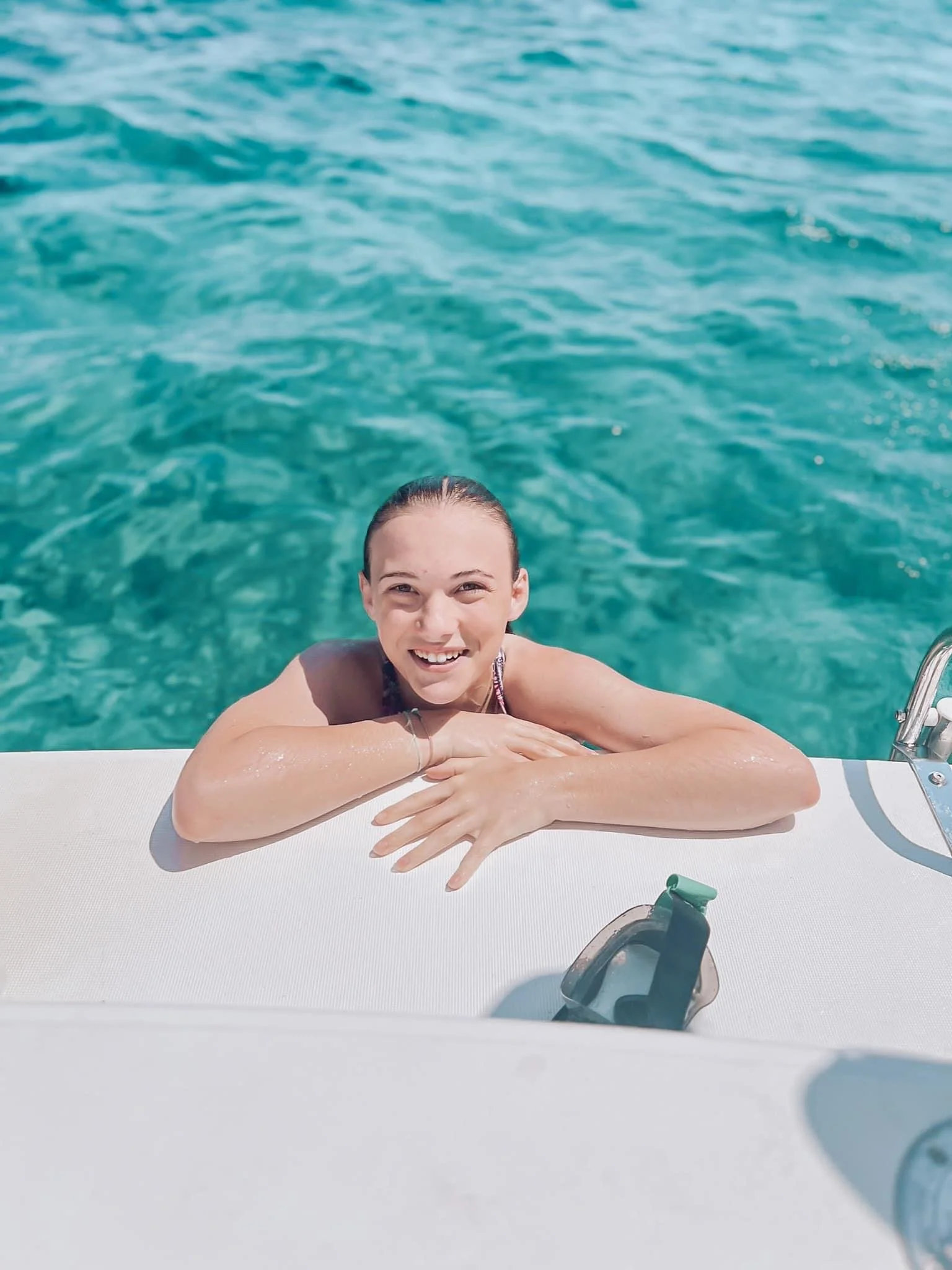 Femme souriante qui sort de l'eau, tenant la main sur le rebord d'un bateau ou d'une plateforme au bord de la mer turquoise, lunettes de soleil posées devant elle.