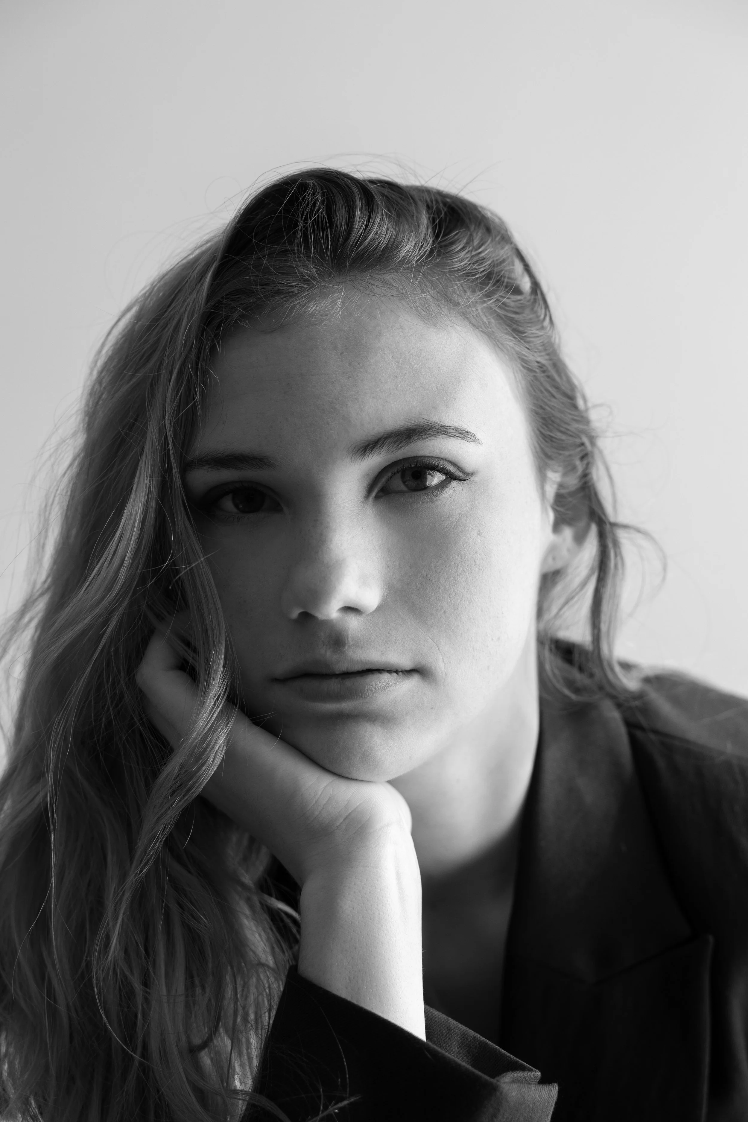 Portrait en noir et blanc d'une jeune femme regardant vers l'objectif, avec ses cheveux longs et ondulés, portant une main sous le visage, vêtue d'un haut sombre.