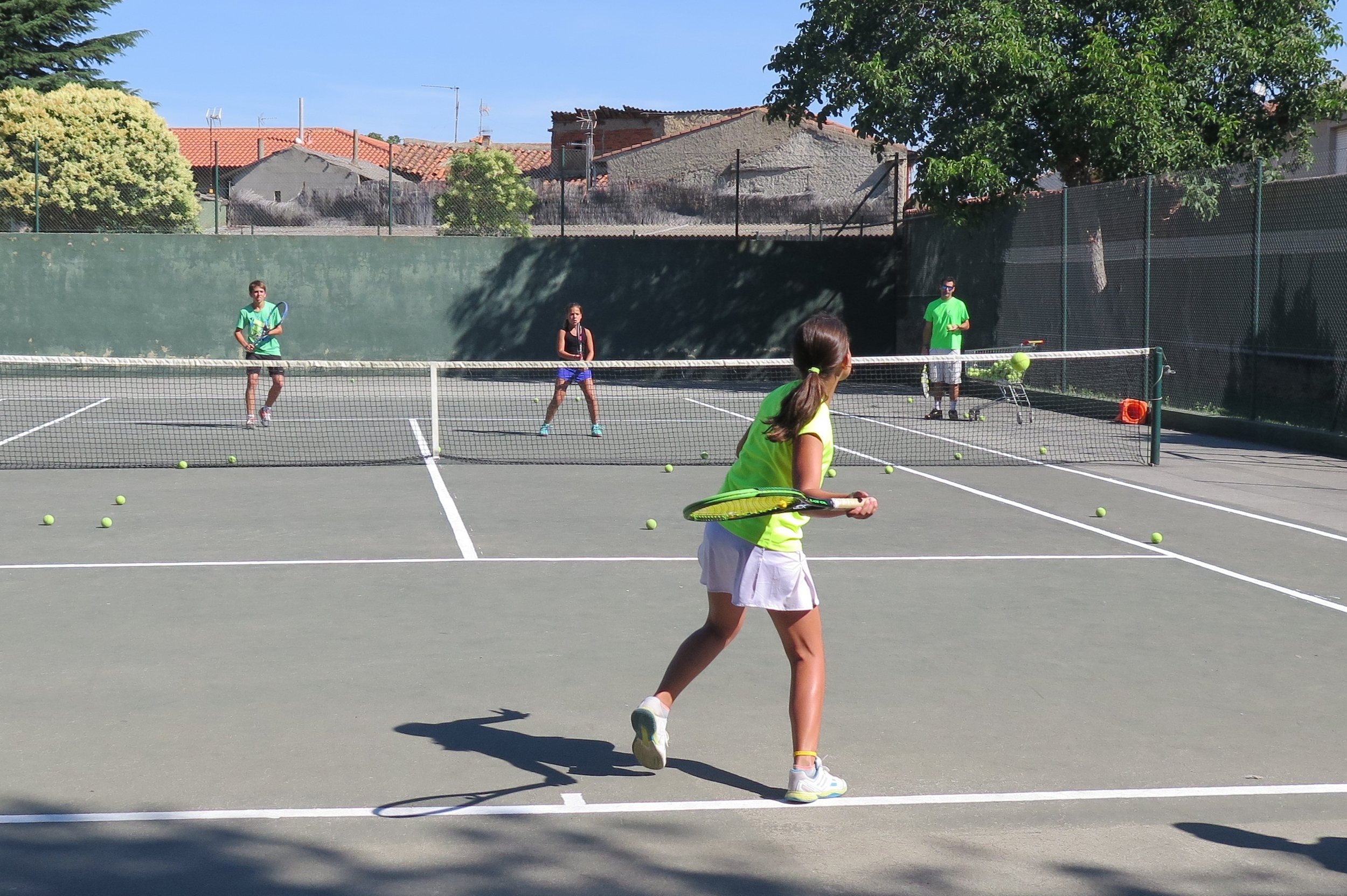 Niños y niñas jugando al tenis en una cancha al aire libre en un día soleado, rodeados de árboles y casas con techos de tejas.