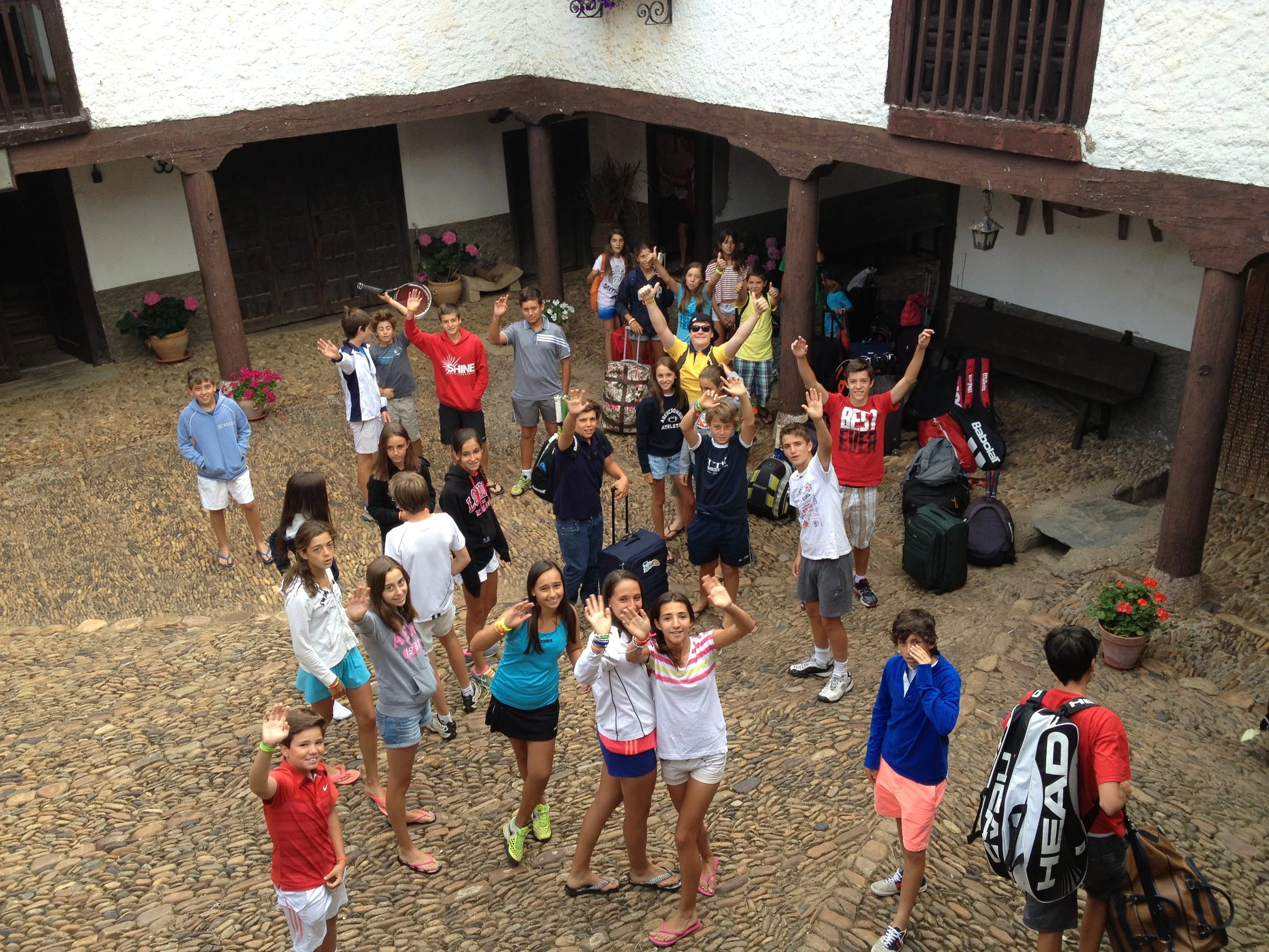 Grupo de niños y adolescentes en un patio de piedra, algunos saludando y otros haciendo gestos, con mochilas y ropa casual. Ambiente alegre y desenfadado.