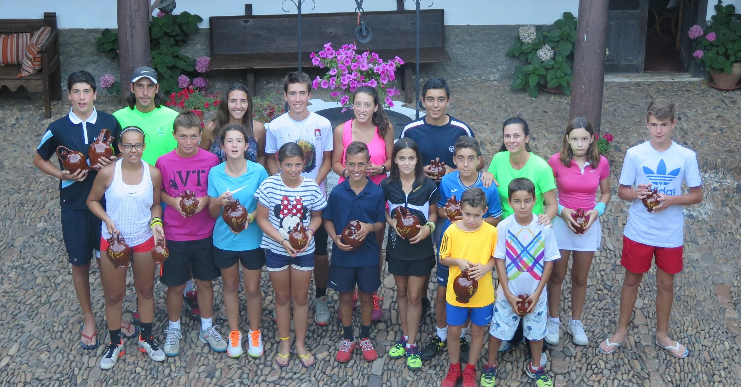 Grupo de niños y adolescentes en un evento al aire libre, sosteniendo cerámicas decorativas, con un fondo de plantas y muebles de madera.