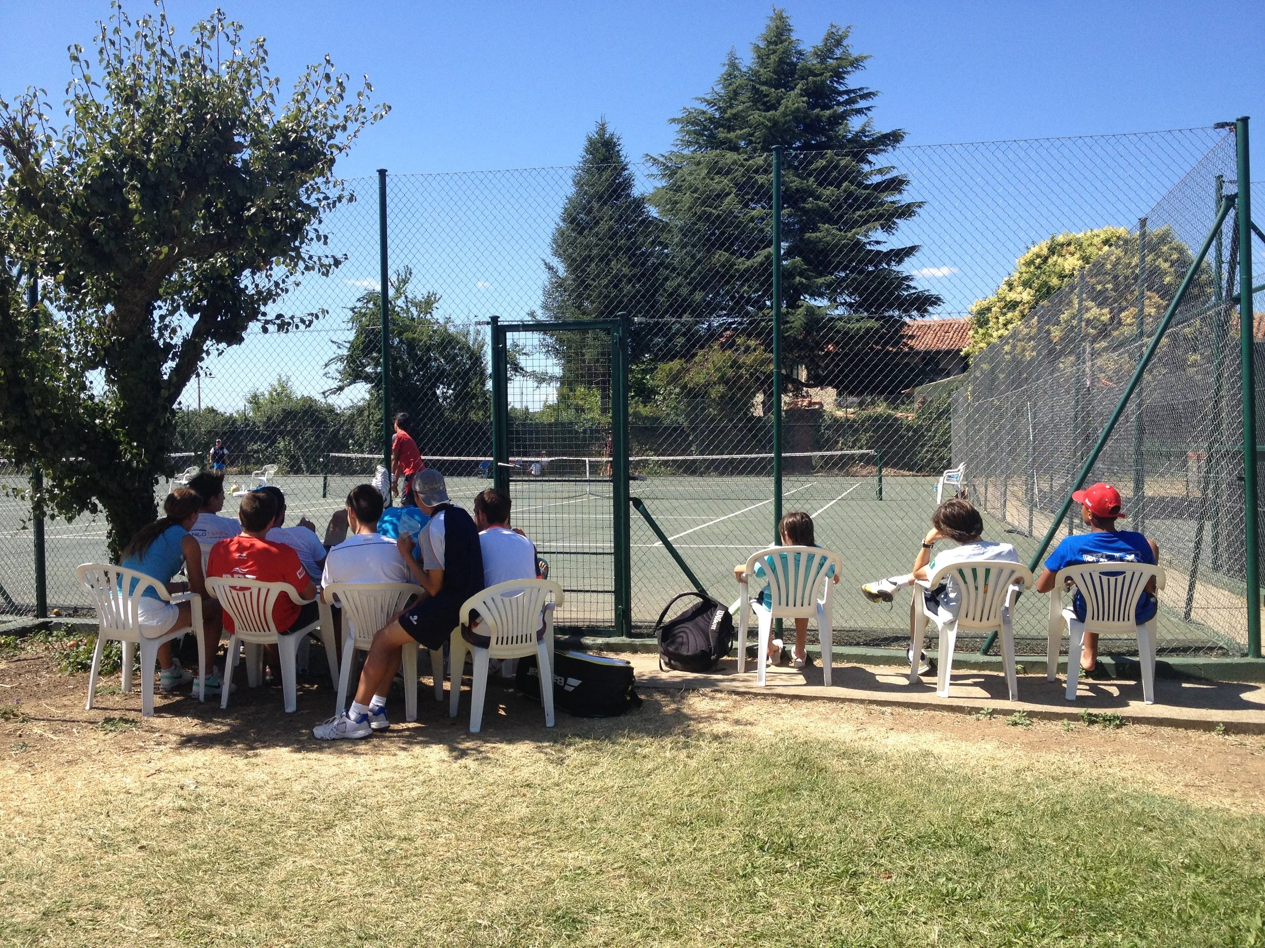Grupo de personas sentadas en sillas plásticas blancas, observando un partido de tenis en una cancha al aire libre, rodeada de árboles y un cielo despejado.