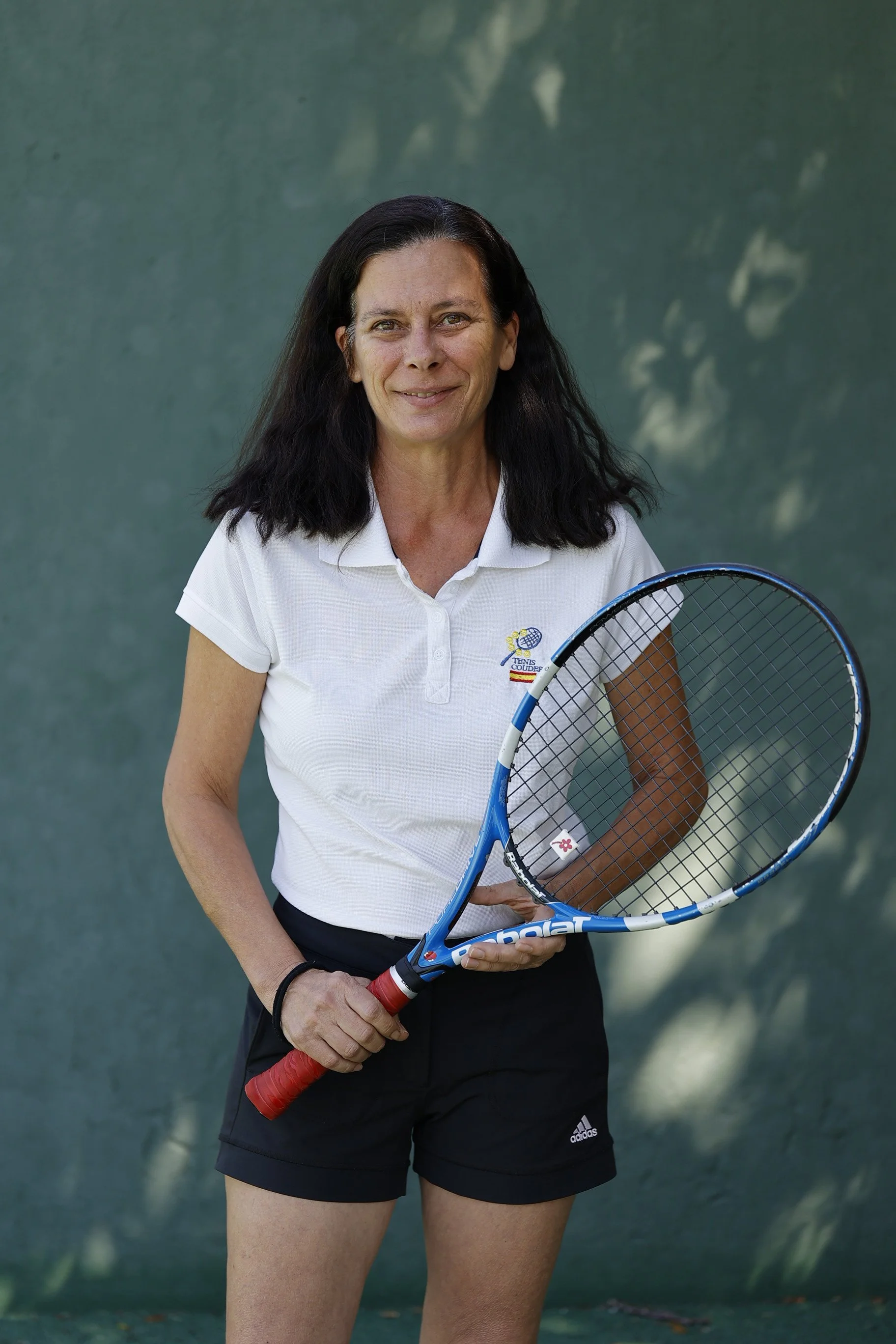 Mujer de cabello largo y oscuro, vistiendo camiseta blanca con logo, pantalones cortos negros, sosteniendo una raqueta de tenis, en una cancha con fondo verde.