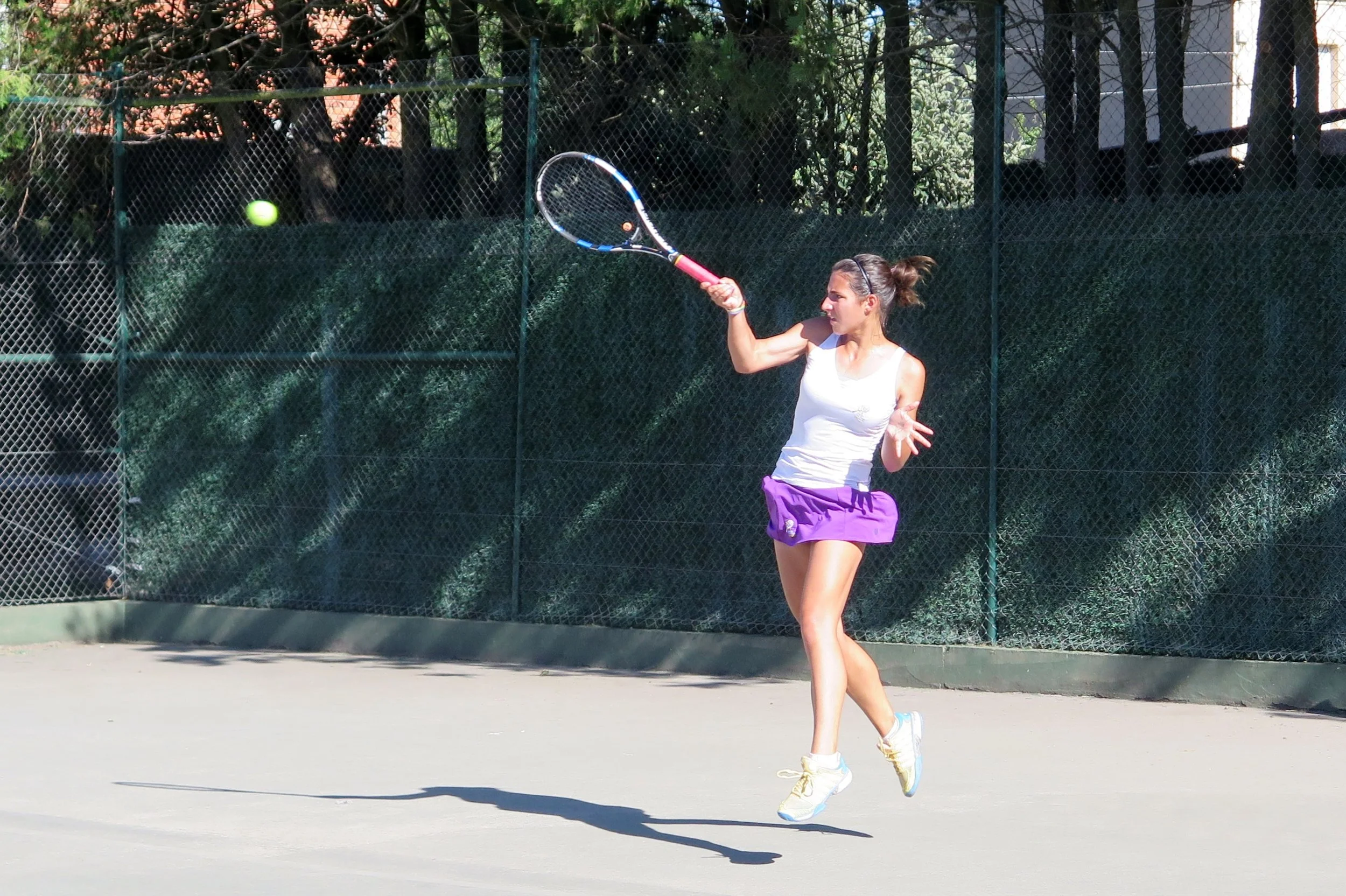 Una mujer jugando tenis en una cancha al aire libre, vistiendo camiseta blanca, falda morada y zapatillas blancas con calcetas amarillas. Ella va a golpear una pelota de tenis con su raqueta.