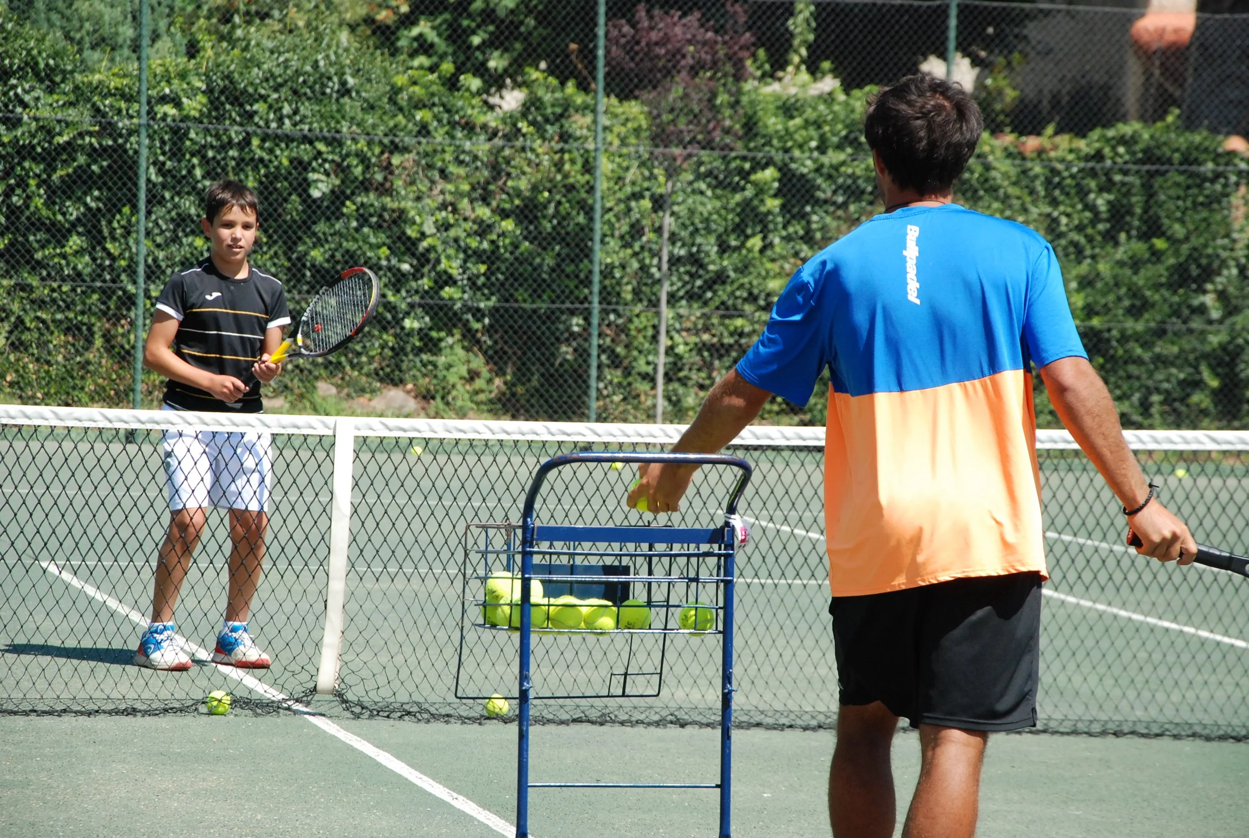 Dos personas jugando tenis en una cancha exterior, con una malla de fondo, una de ellas sosteniendo una raqueta y la otra con una pelotera, varias pelotas en una cesta y en el suelo.