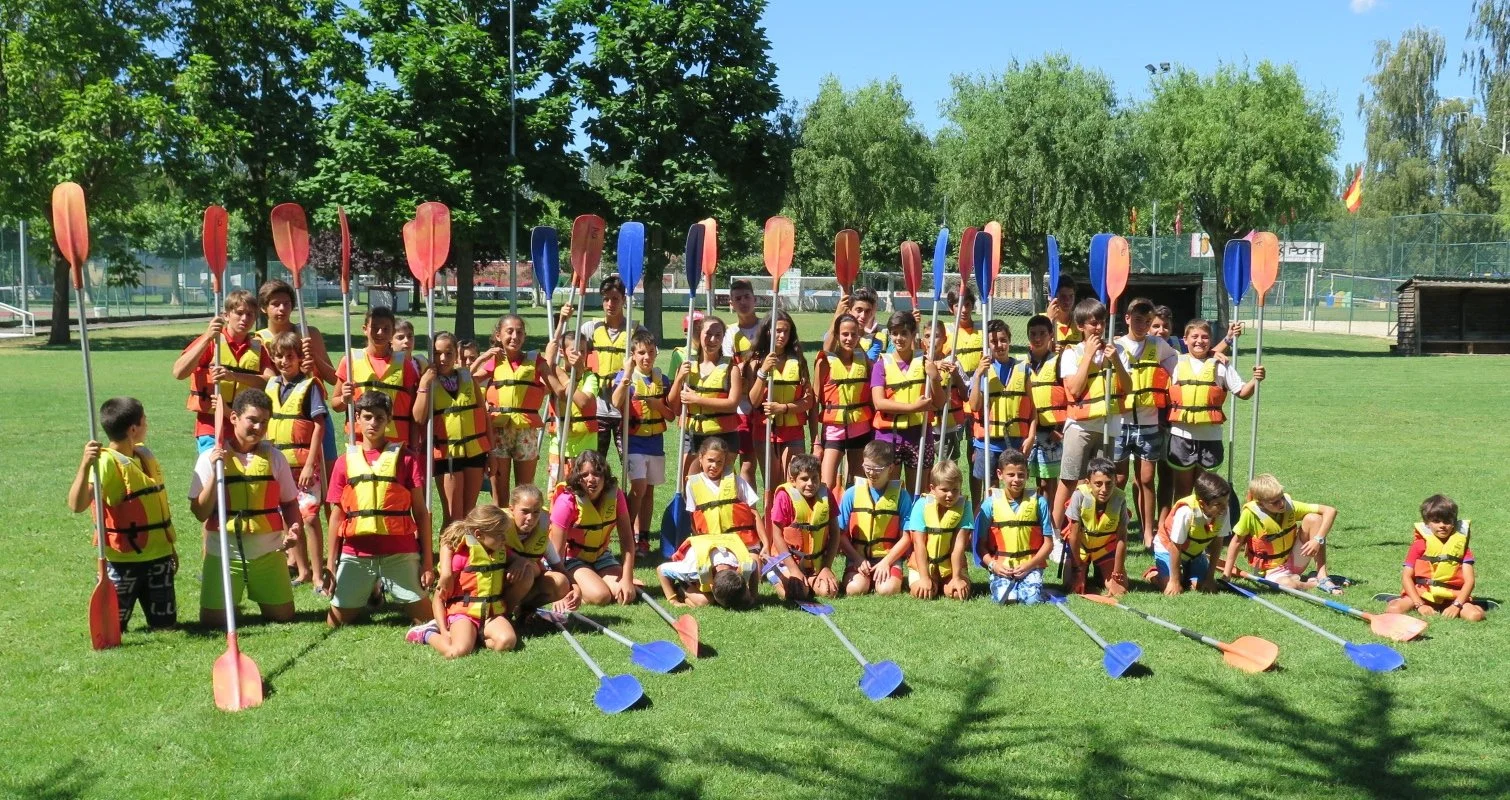 Grupo de niños y niñas con chalecos salvavidas y remos en un campo de césped, listos para una actividad acuática al aire libre, con árboles y una pista de tenis en el fondo, en un día soleado.