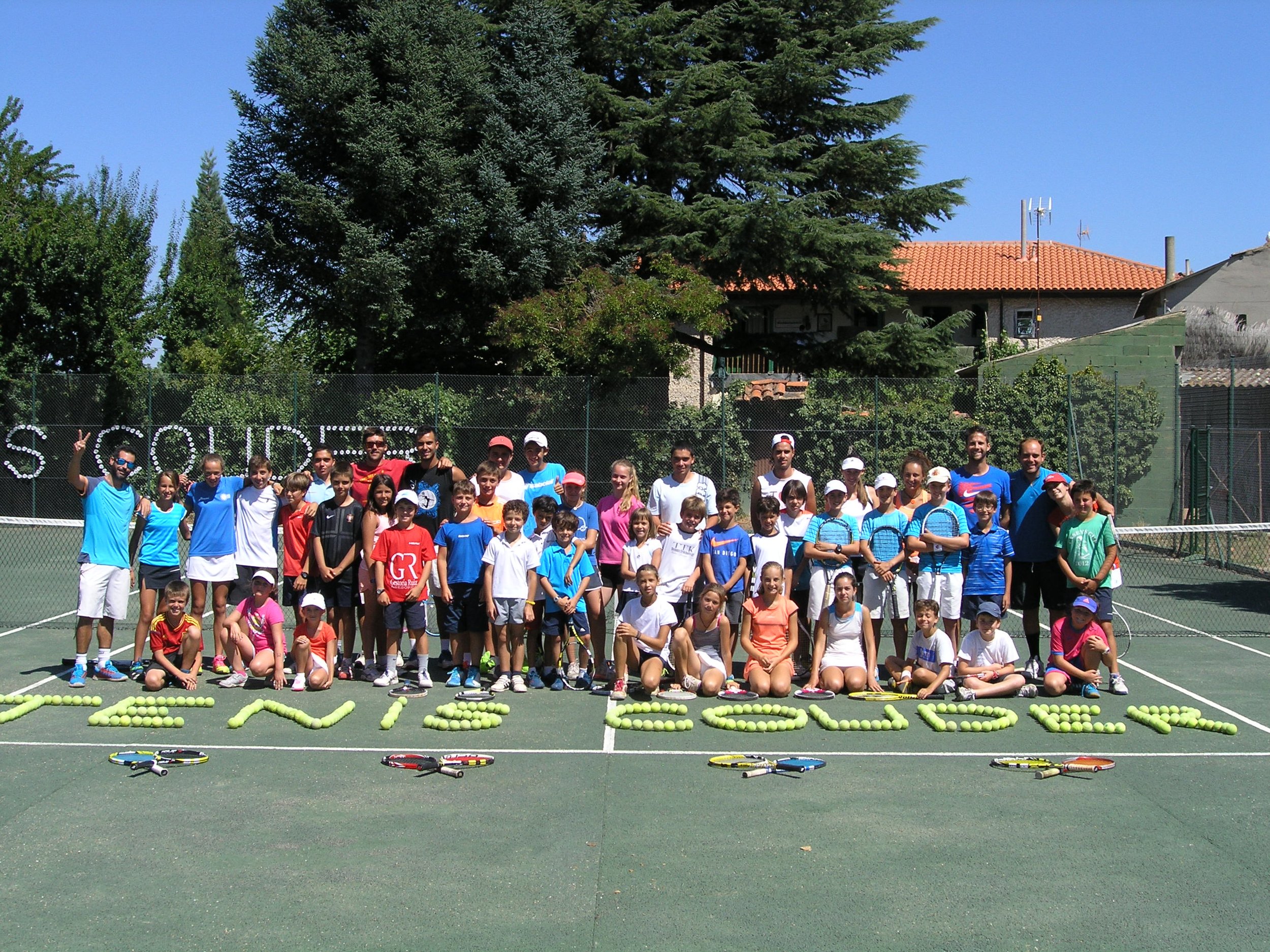 Grupo de niños y adultos en una cancha de tenis, algunos con raquetas, formando la palabra 'EXITO' con pelotas de tenis en el suelo y sosteniendo una pancarta que dice 'SCHOOL' al fondo. Se observa un árbol grande y una casa con techo rojo en el fondo.