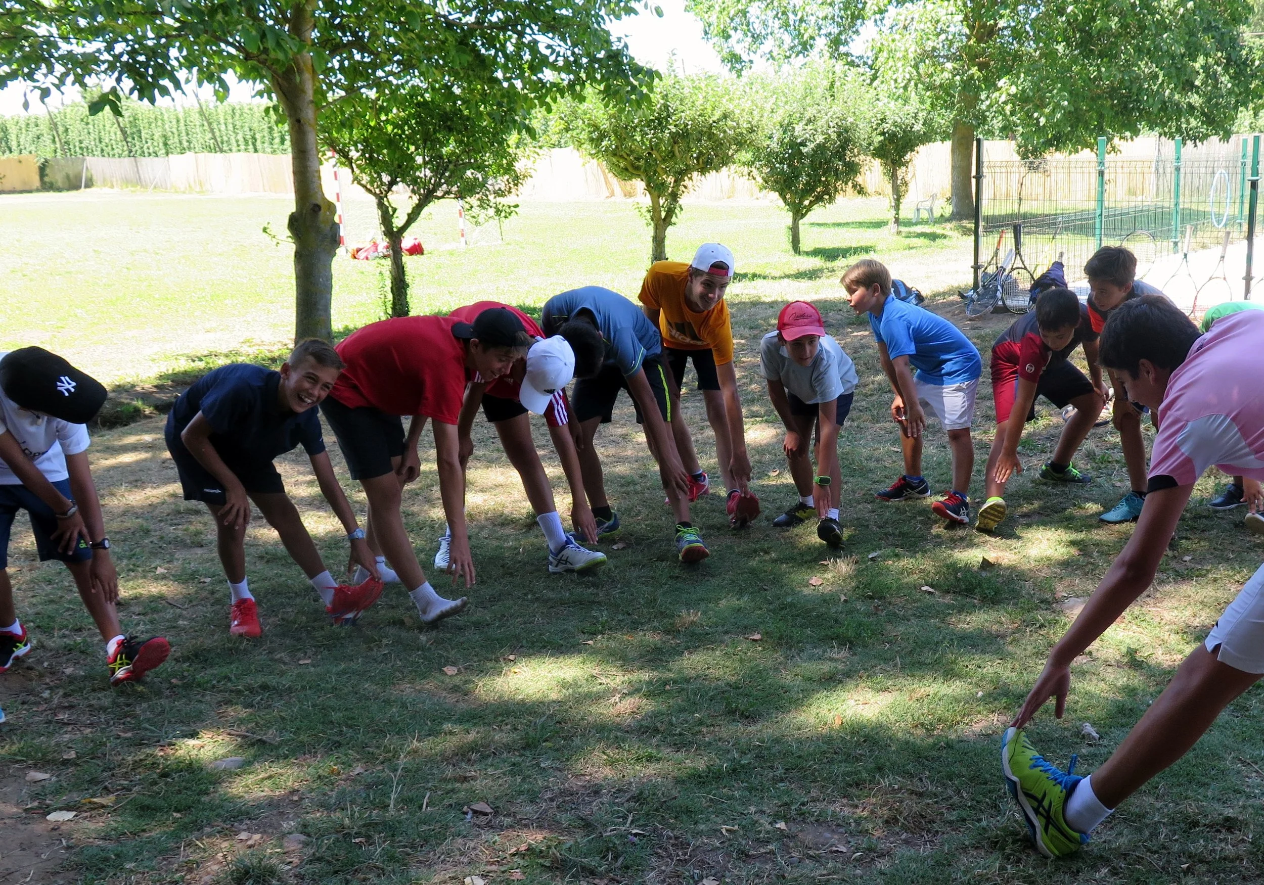 Grupo de niños participando en una actividad al aire libre, agachados con las manos en las rodillas, en un parque soleado con árboles y un fondo de campo con enrejado y una portería.