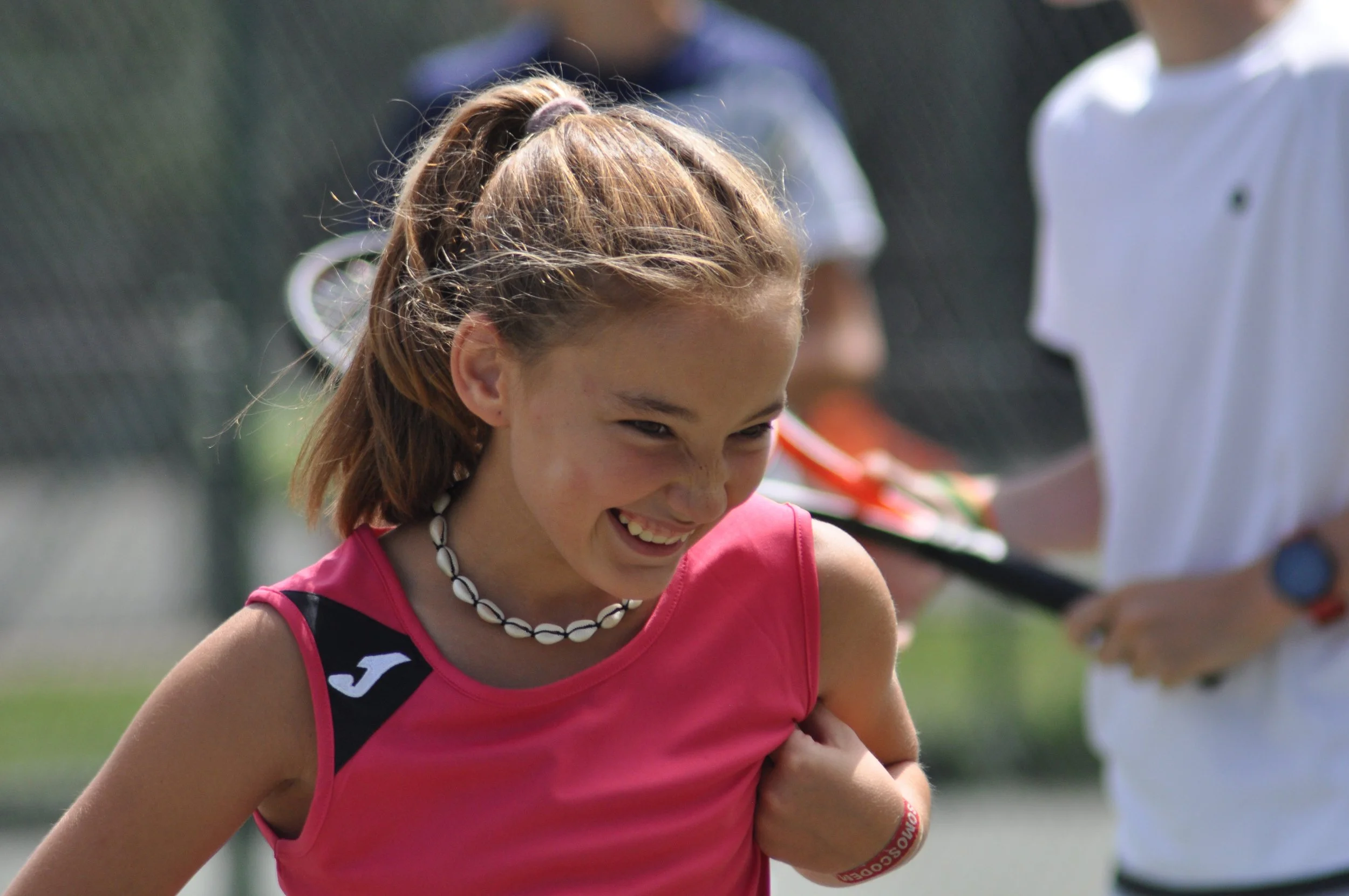 Una niña sonriendo, vestida con ropa de deporte rosa y un collar de conchas, participando en un evento al aire libre con varias personas en el fondo.
