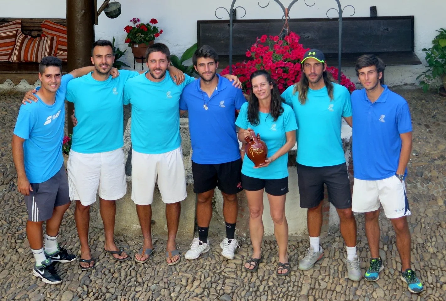 Grupo de siete personas, seis hombres y una mujer, posando juntos en un entorno exterior. Todos llevan ropa deportiva de color azul. La mujer sostiene una vasija de cerámica marrón. Detrás hay plantas, flores rojas y un banco con cojines. La superfic