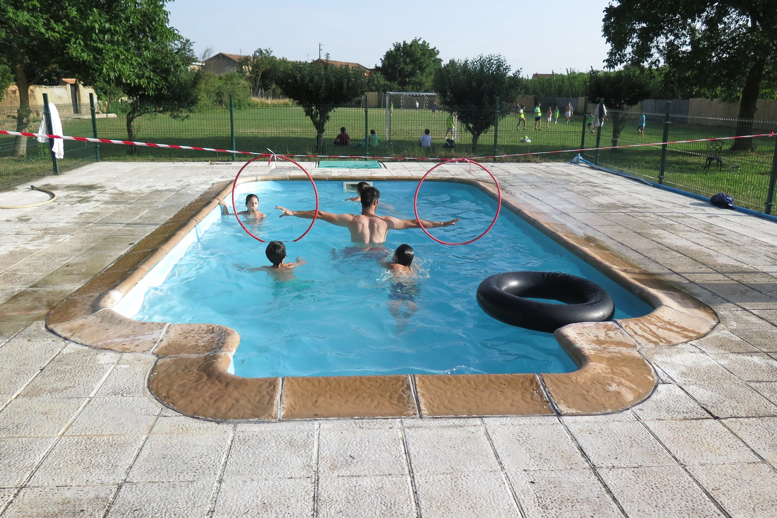 Niños y un instructor jugando en una piscina en un entorno al aire libre, con un campo de fútbol y árboles en el fondo.