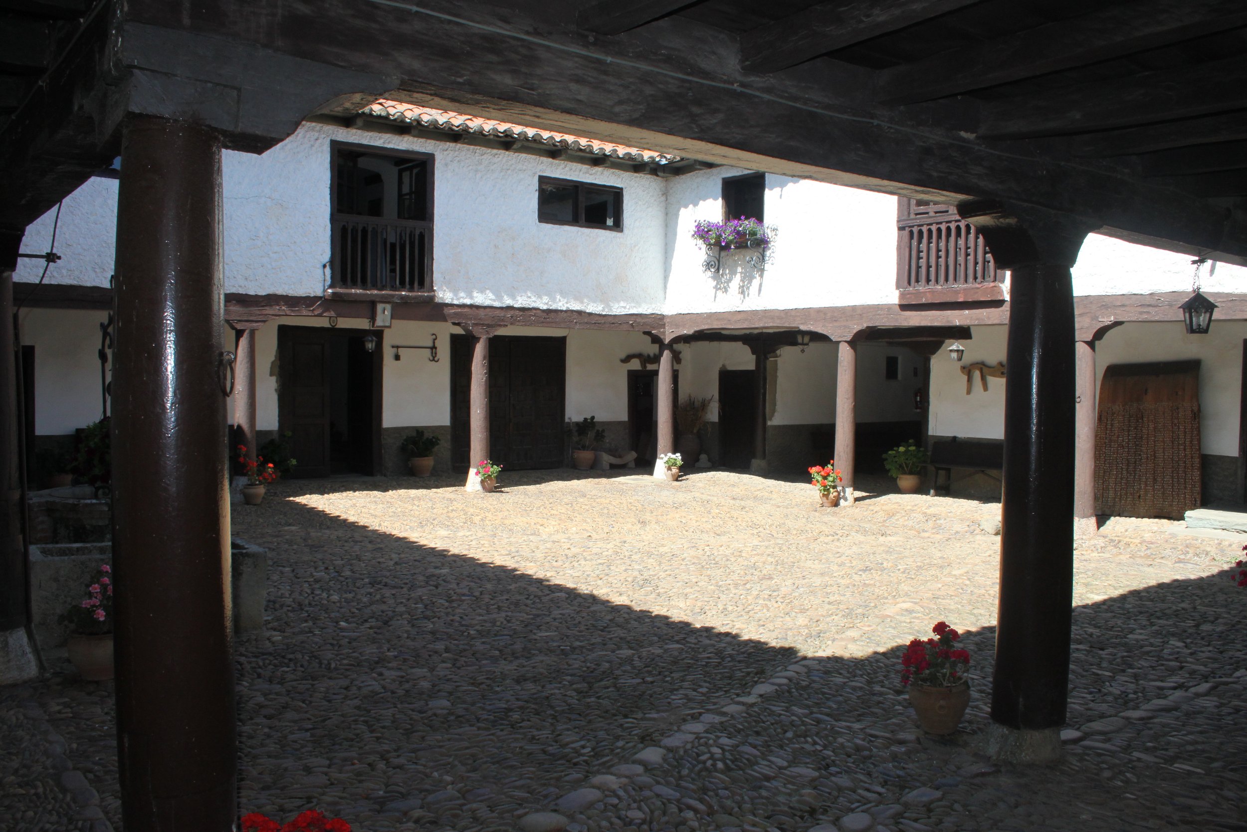 Vista de un patio de casa colonial con pilares de madera y paredes blancas, decorado con macetas de flores y ventanas con rejas de hierro.