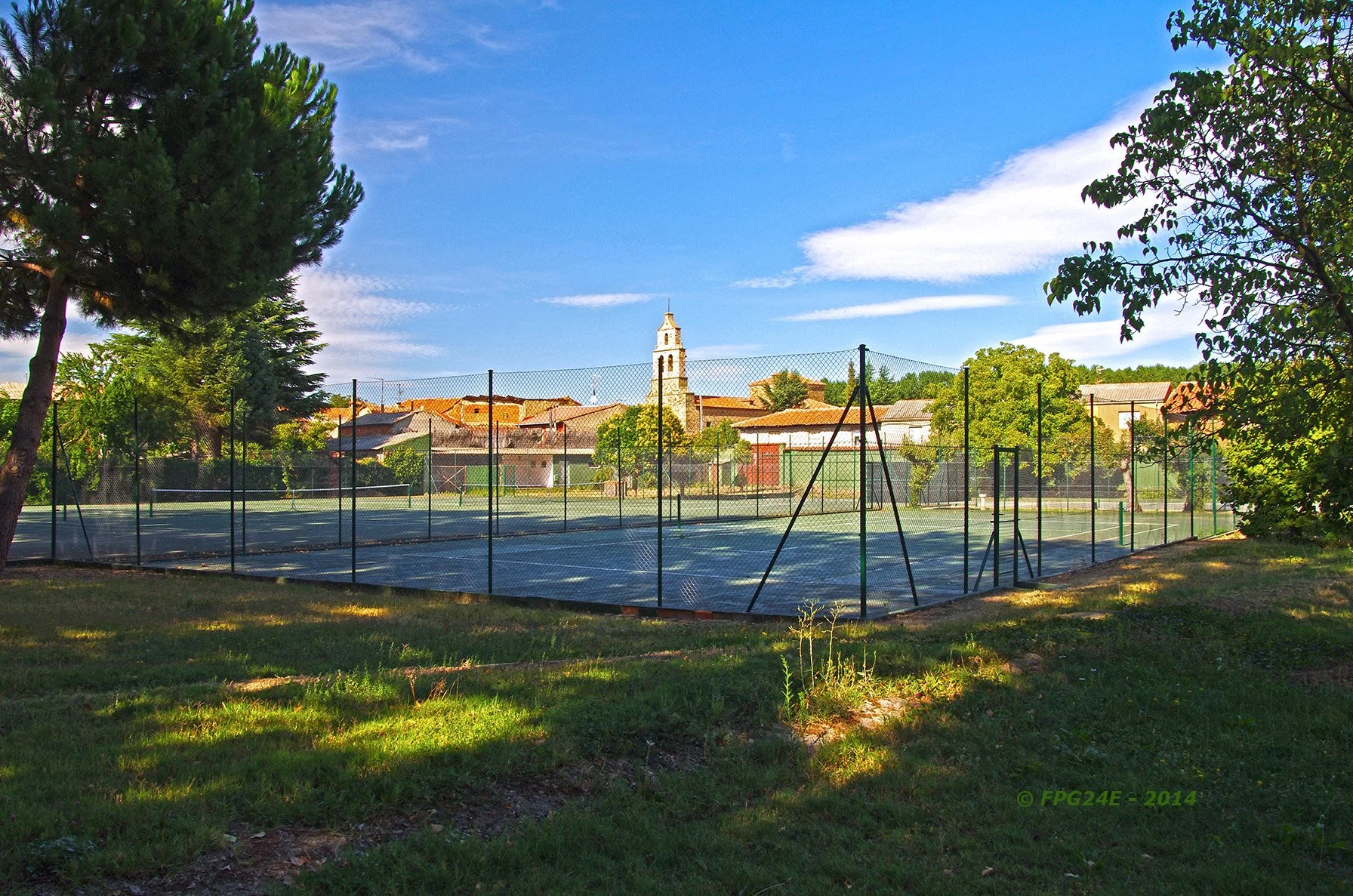 Cancha de tenis rodeada de árboles y cercas con un pueblo y una iglesia en el fondo, en un día soleado con cielo despejado.