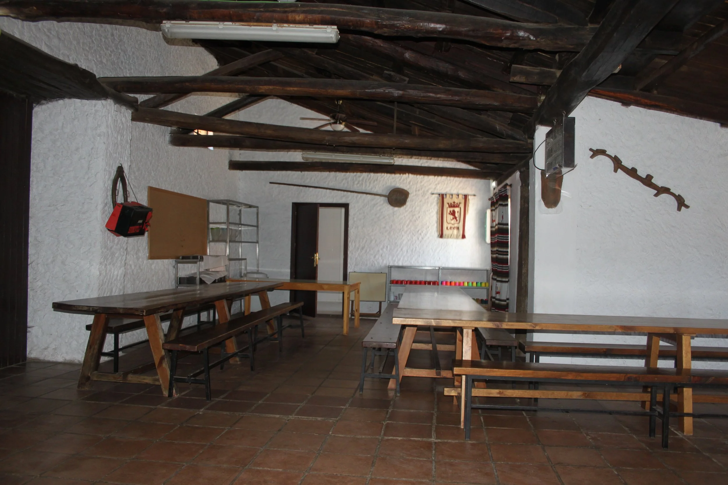 Una habitación con paredes blancas y techo de madera, con mesas y bancos de madera, un ventilador de techo, una estantería metálica, y decoraciones tradicionales en la pared.
