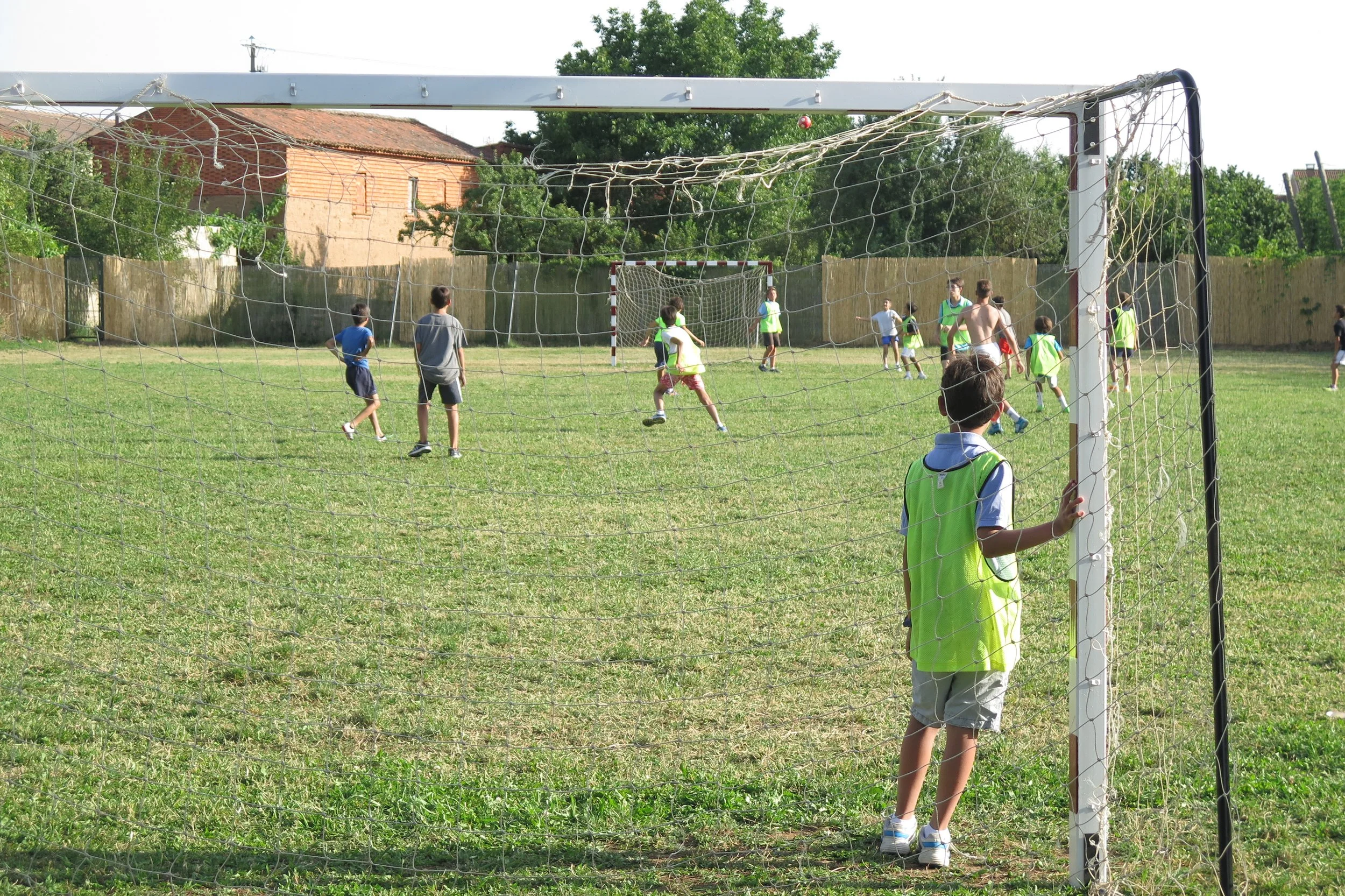 Niños jugando fútbol en un campo abierto, con algunos niños con camisetas verdes y otros con camisetas blancas, mientras un niño observa desde el borde del campo.