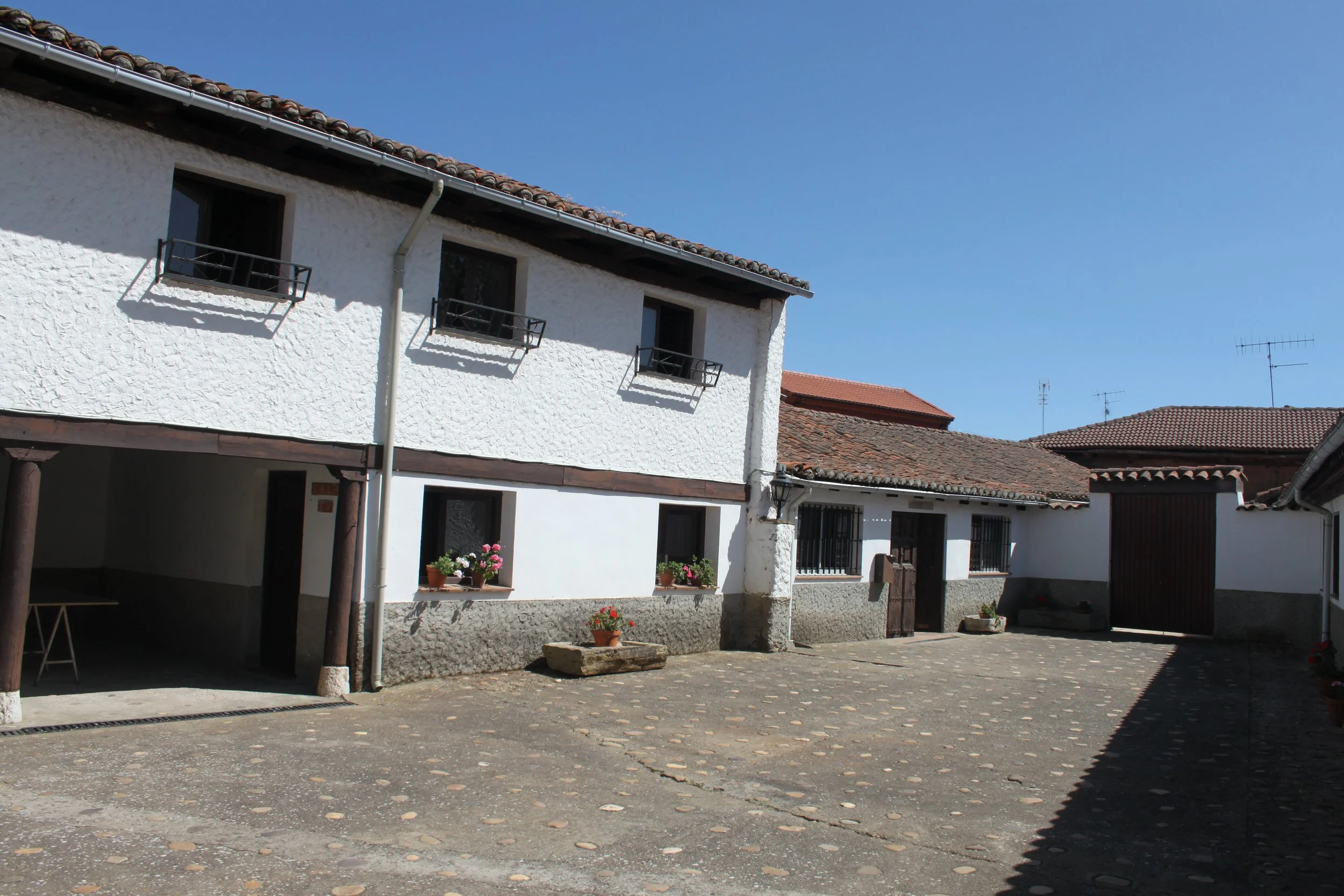 Vista de una casa de paredes blancas y techo de tejas rojas, con pequeñas ventanas con barrotes y balcones. Hay algunas plantas con flores en la entrada y un espacio abierto con piso de piedra.