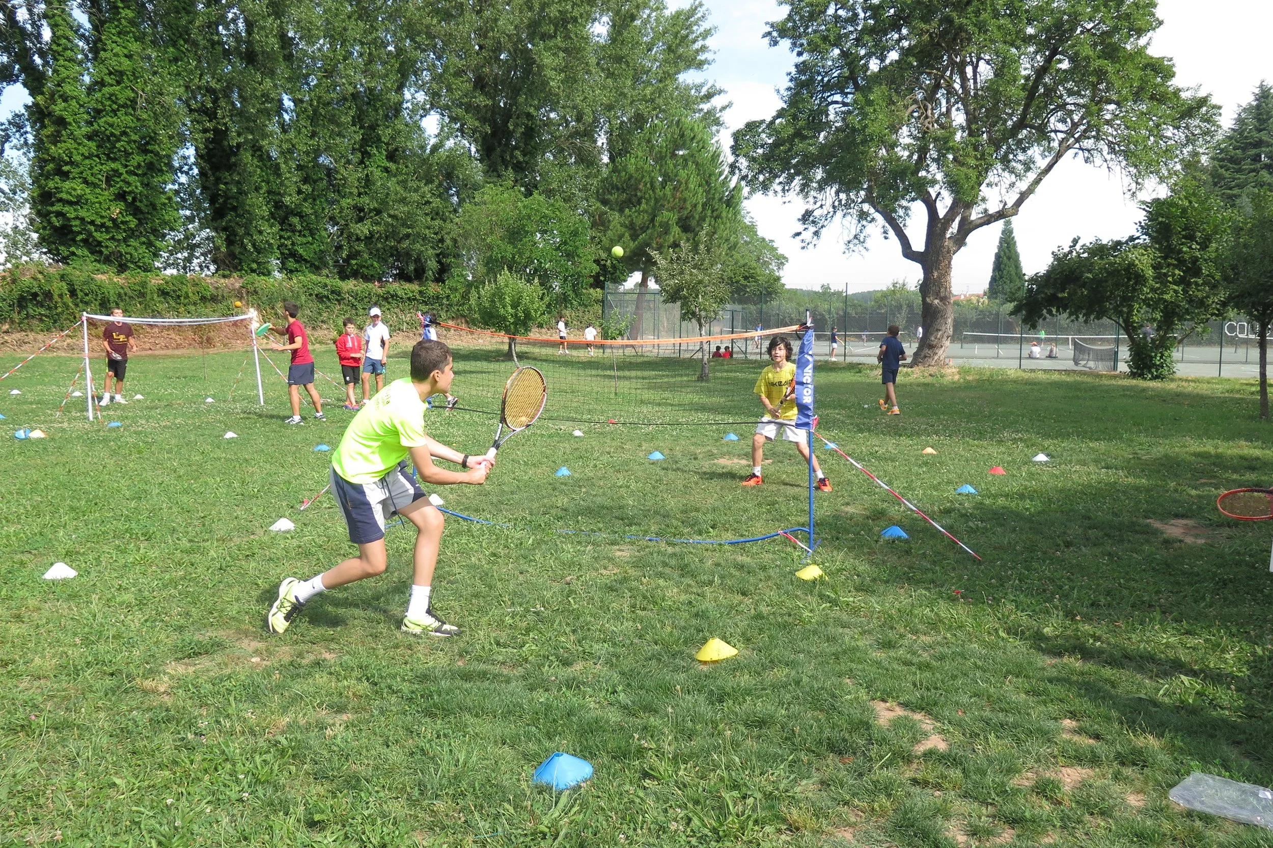 Niños jugando al tenis en un parque, participando en actividades de entrenamiento con conos y redes, rodeados de árboles y césped.