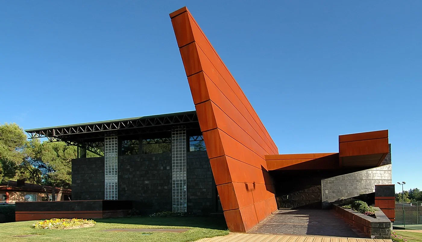 Edificio moderno con estructura de metal y madera en forma angular, rodeado de césped y plantas, bajo un cielo despejado.