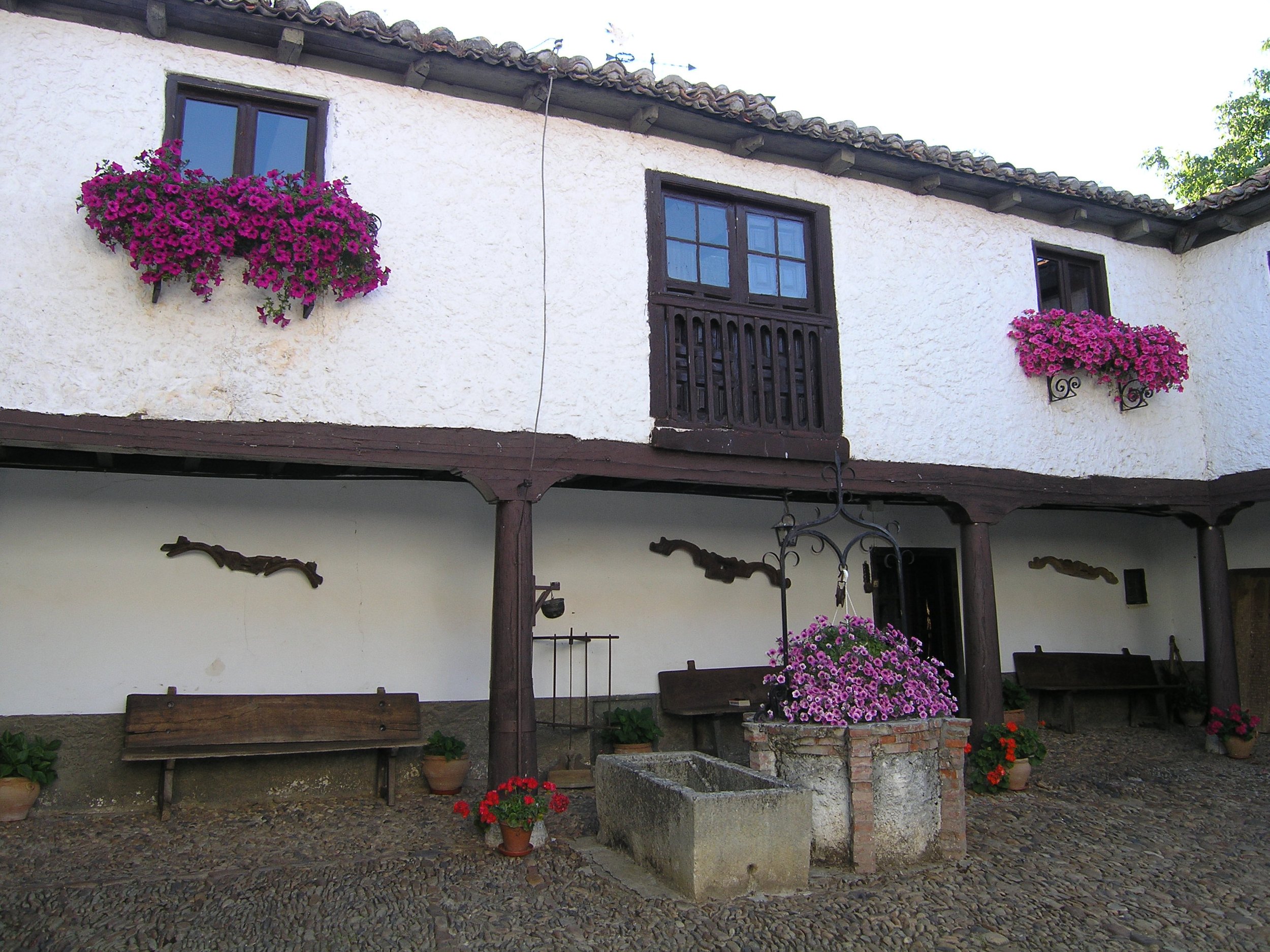 Casa tradicional con paredes blancas, ventanas con marcos de madera y macetas con flores rosa y moradas colgadas en la fachada. Debajo hay bancos, flores en macetas y un pozo de piedra.
