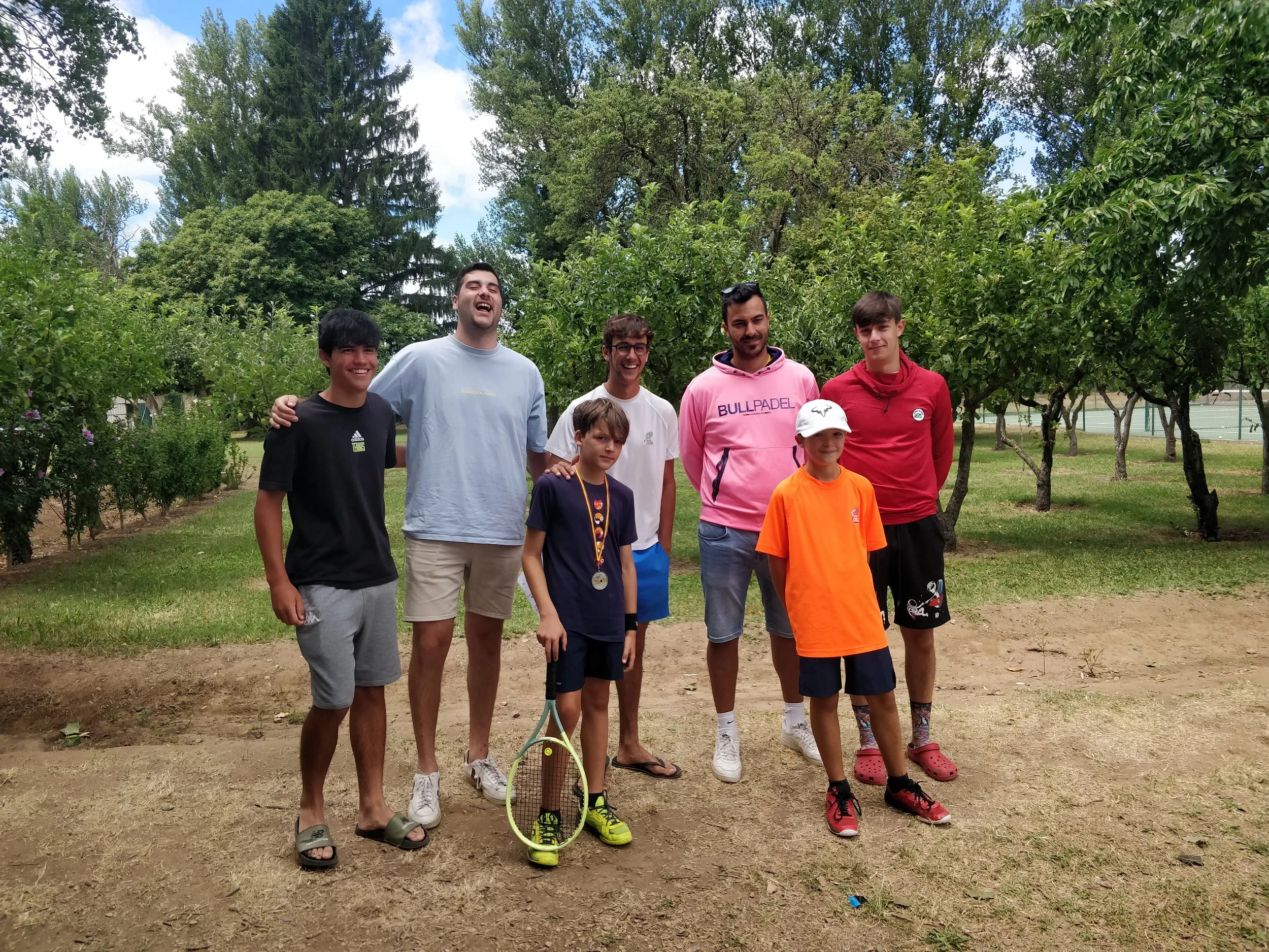 Grupo de siete personas jóvenes sonriendo y posando al aire libre en un parque con árboles y cancha de tenis al fondo, algunos llevan ropa deportiva y uno sostiene una raqueta de tenis.