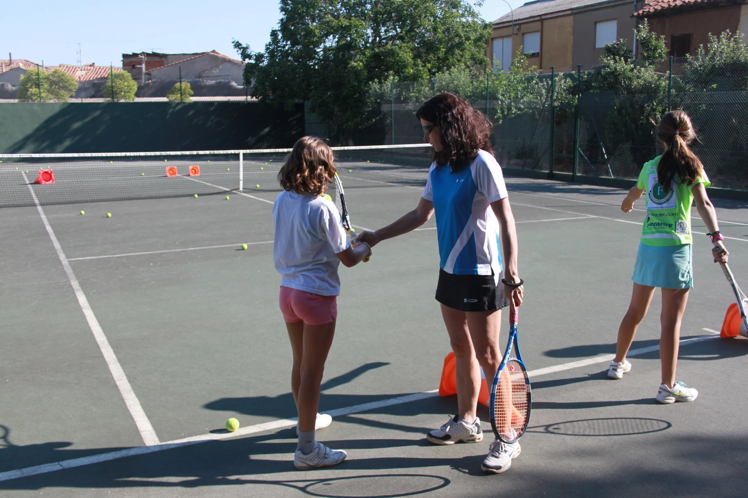 Tres niñas y una mujer en una cancha de tenis, con varias pelotas de tenis y conos de entrenamiento, participando en una actividad de tenis al aire libre.