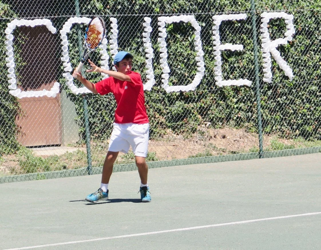 Un niño jugando al tenis en una cancha exterior, vestido con camiseta roja, pantalones cortos blancos y gorra azul, sosteniendo una raqueta.