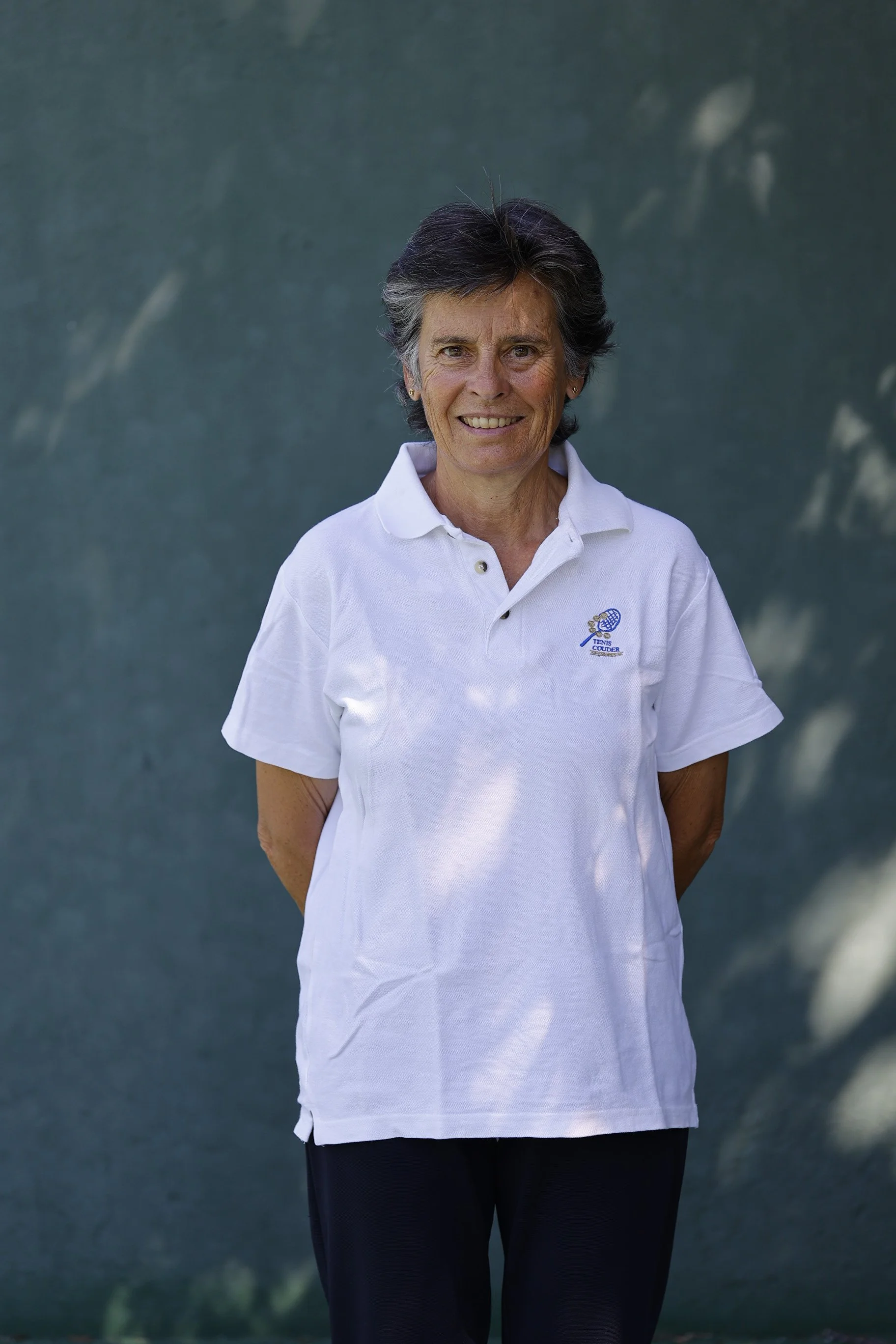 Mujer mayor sonriendo con camisa blanca con logotipo de tenis, de pie al aire libre frente a un fondo difuso y oscuro.