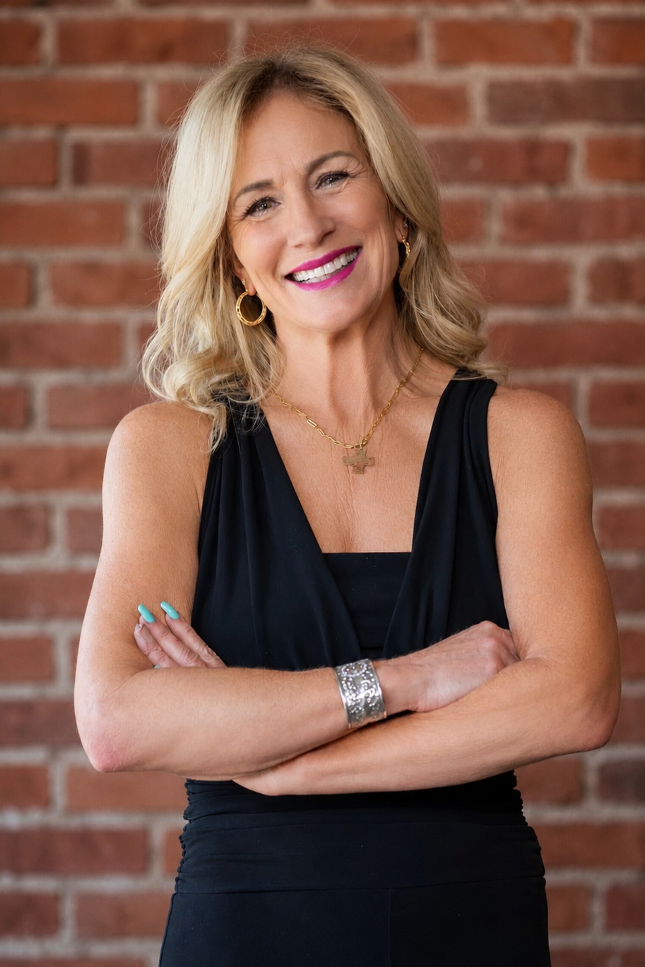 A smiling middle-aged woman with blonde, wavy hair and wearing a black sleeveless dress, accessorized with gold hoop earrings, a gold necklace, a silver bracelet, and bright pink lipstick, standing against a brick wall background.