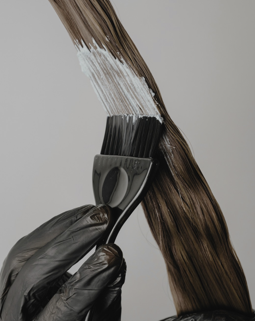 Close-up of a hand wearing a black glove holding a paintbrush, applying white paint to a strand of brown hair against a plain light background.