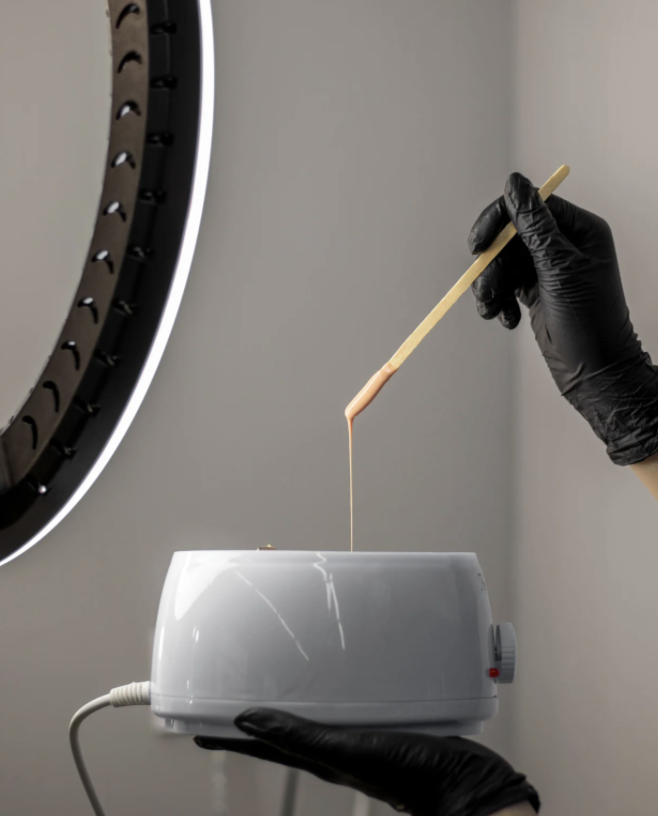 A person wearing black gloves uses a wooden stick to dip into a small white wax warmer as melted wax drips from the stick.