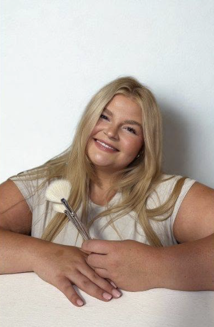 Young woman with long blonde hair smiling at the camera, holding makeup brushes, sitting at a table against a plain white wall.