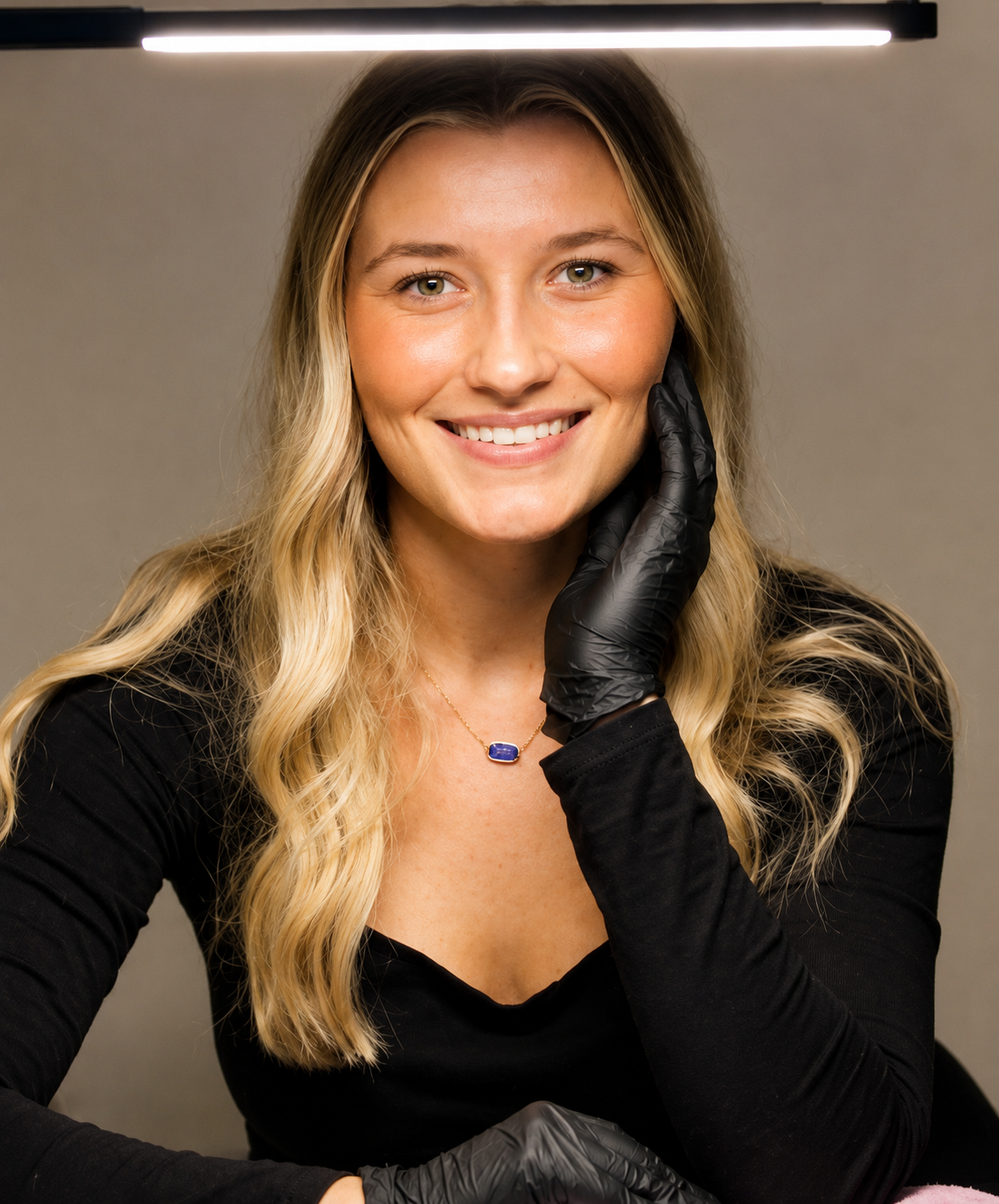 A woman with long blonde hair wearing a black long-sleeve top and black gloves, smiling and touching her face with her right hand, in front of gray background.