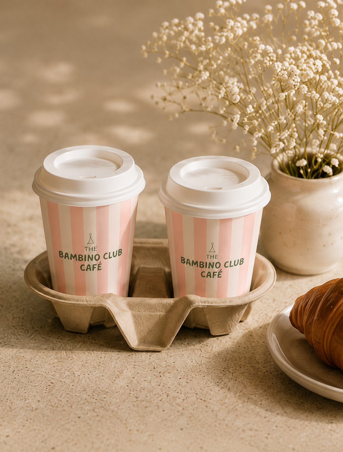 Two pink and white striped coffee cups with white lids labeled 'The Bambino Club Café,' placed in a biodegradable tray on a beige countertop. A vase with white baby's breath flowers and a plate with a croissant are nearby.