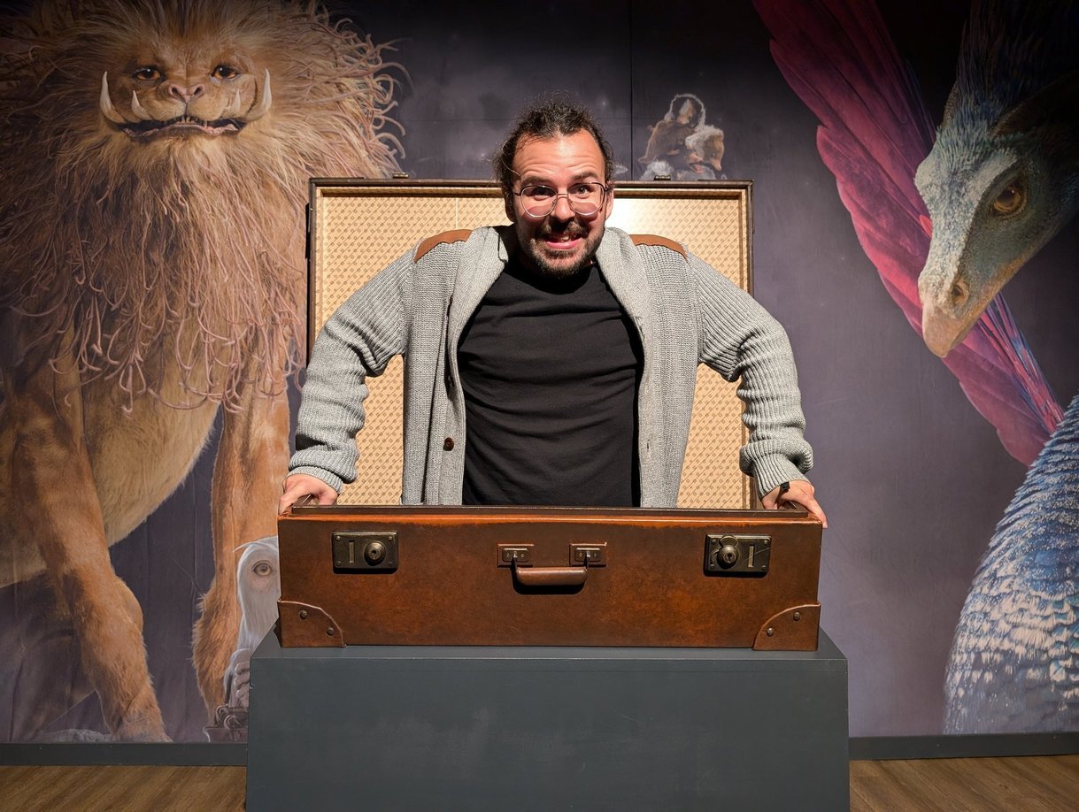 Un homme souriant avec des lunettes, debout derrière un coffre en bois, placé sur une table. Il y a un décor mural avec une lionne, un perroquet et un pangolin