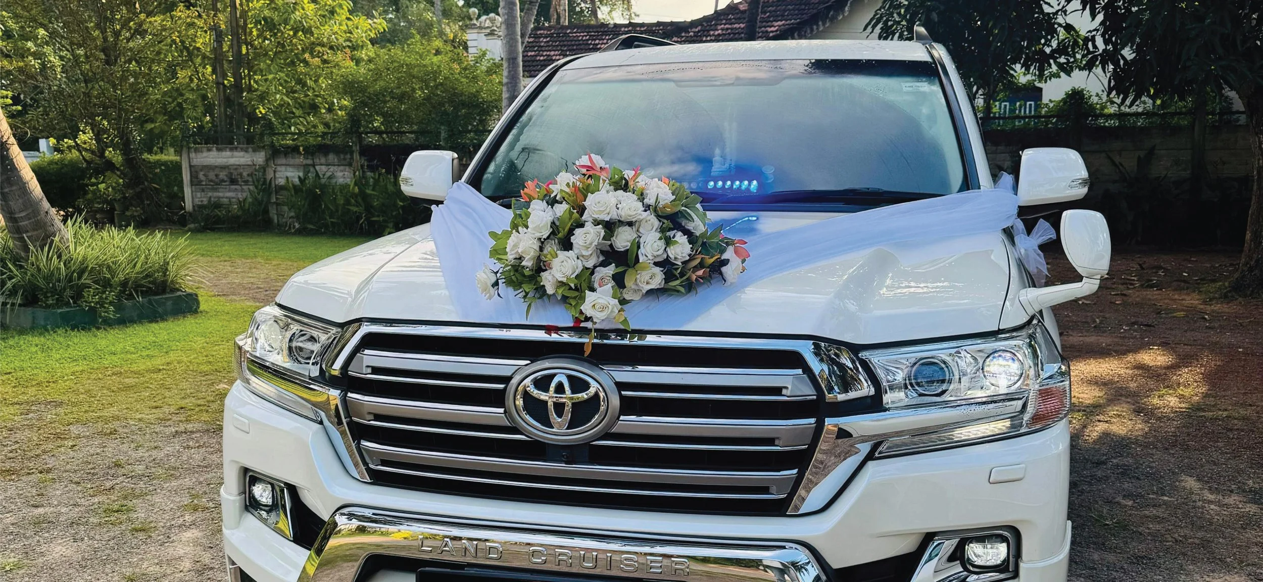 White Toyota Land Cruiser decorated with a white ribbon and a bouquet of white roses and pink flowers for a wedding or special event, parked outdoors in a green garden.