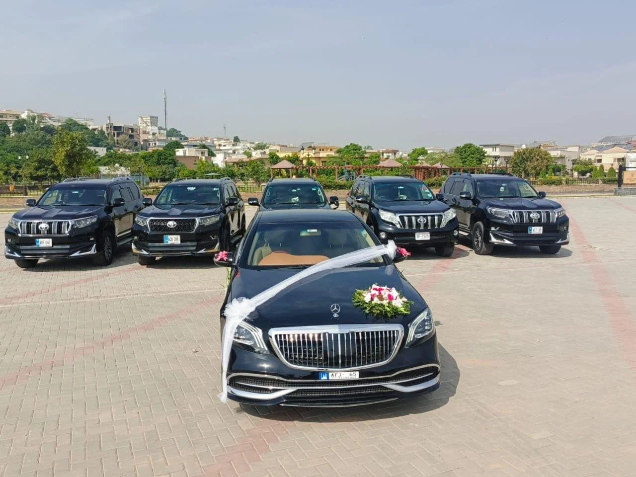 Black Mercedes-Benz with wedding decorations parked in front of five black Toyota SUVs on a spacious paved lot, with a residential area in the background.