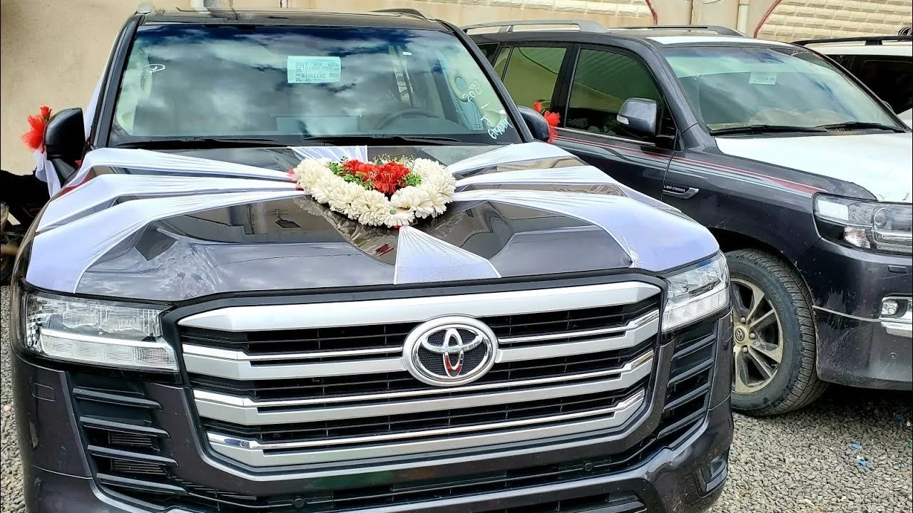 Black Toyota SUV decorated with a floral bride and groom topper, ribbons, and flowers, parked next to a black SUV on a gravel lot