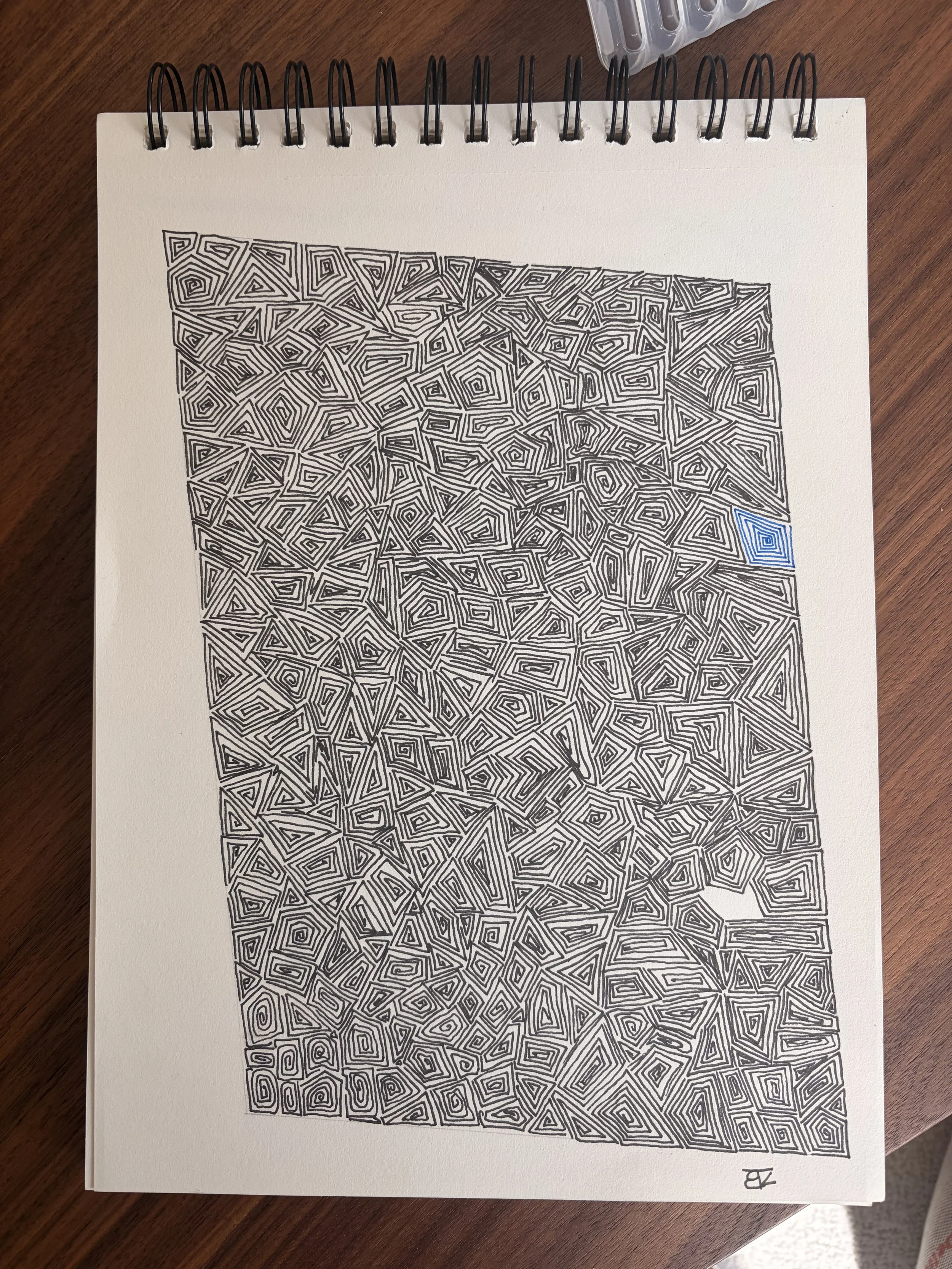 A black and white abstract geometric drawing on a spiral notebook page, containing intricate maze-like patterns and a blue square in the bottom right corner. The notebook is placed on a wooden surface.