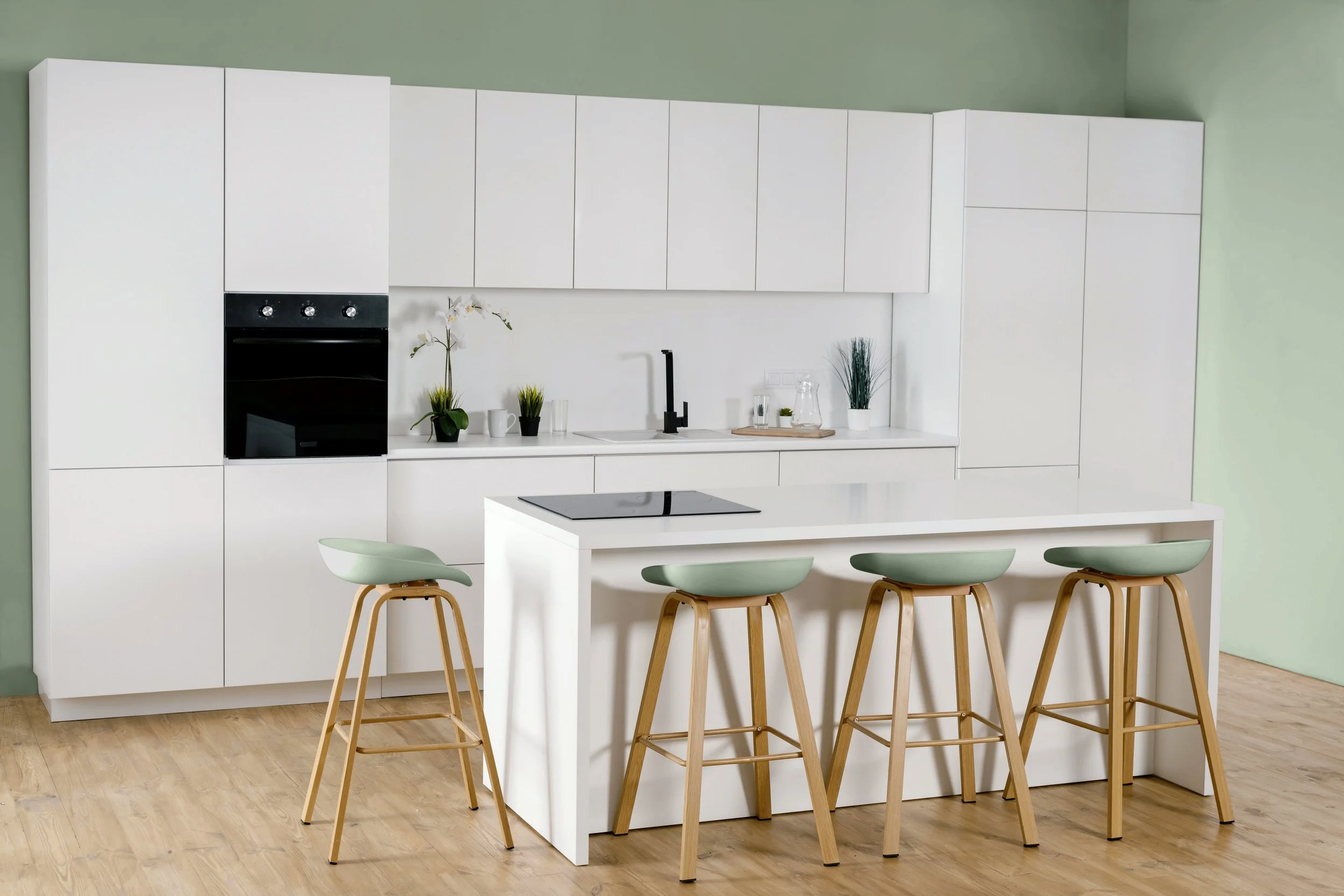 Cuisine moderne avec armoires blanches, un îlot central blanc, chaises en bois avec sièges en vert pastel, décor minimaliste avec plantes et accessoires