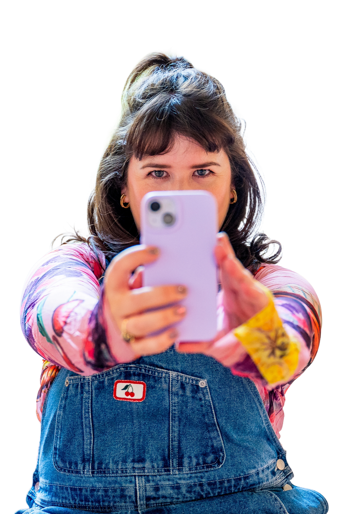 A young woman with brown hair and blue eyes taking a selfie with a pink smartphone, wearing a colorful patterned shirt and denim overalls with a cherry patch, sitting down with her legs crossed.